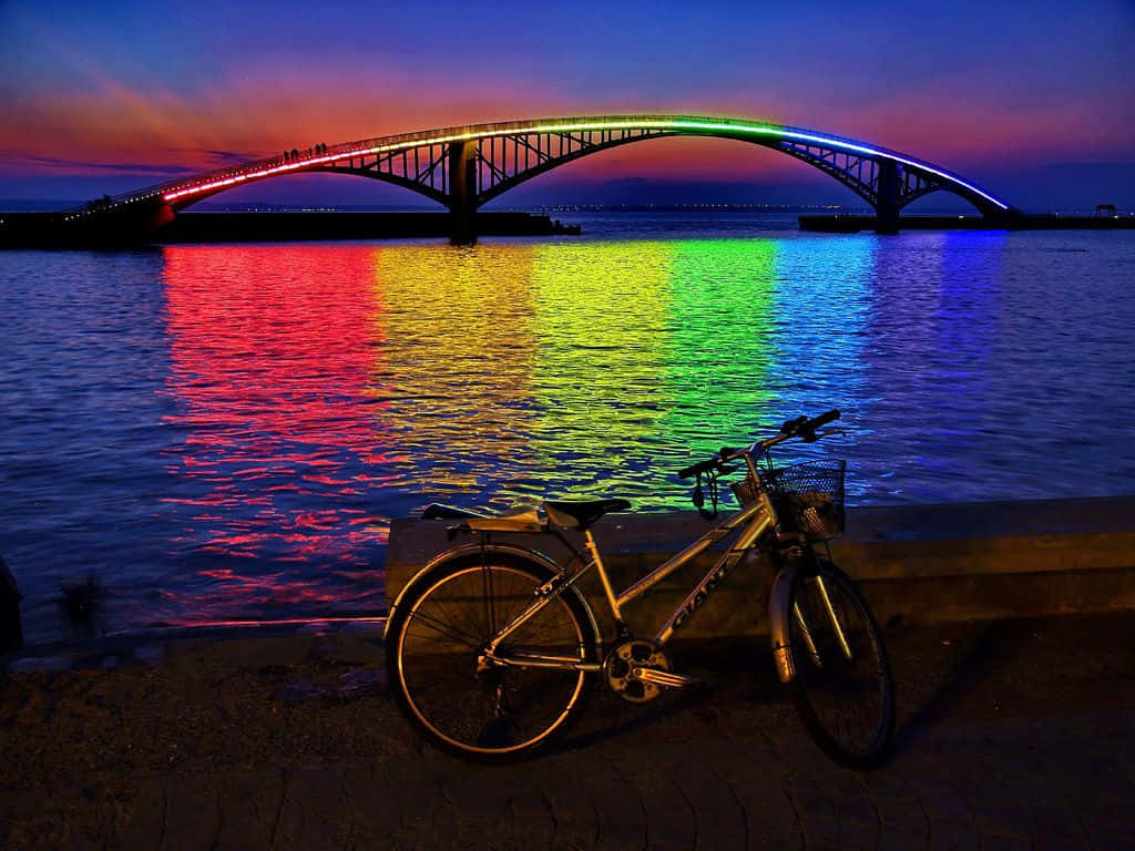 Rainbow Bridge 桌布 壁紙 — 一輛腳踏車停在水邊，彩虹燈光下的橋樑在暮色波浪中倒映。