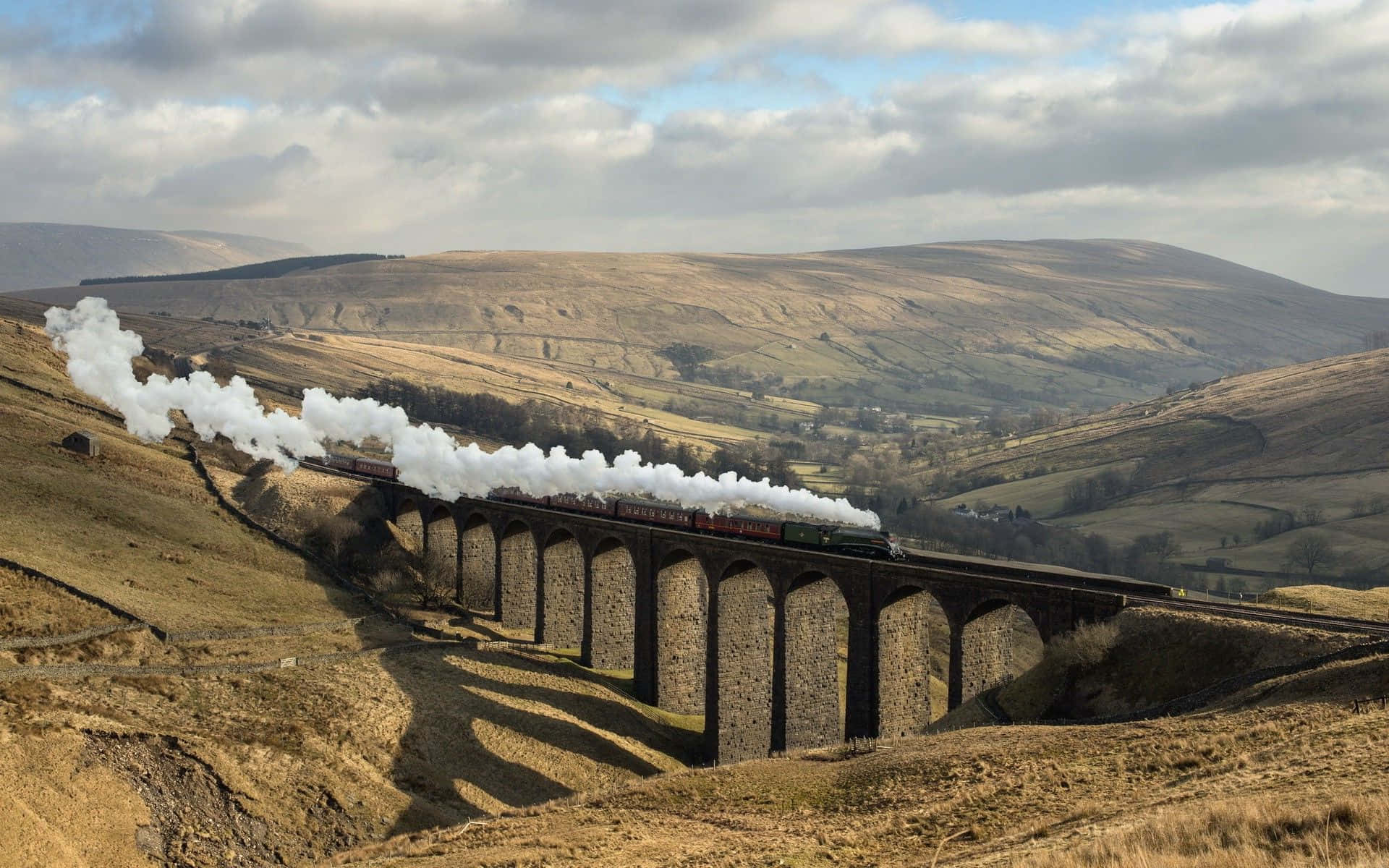 Hogwarts Express 桌布 壁紙 — 一輛蒸汽火車噴出白煙，穿過多雲天空下起伏的金色山丘中的石拱橋。