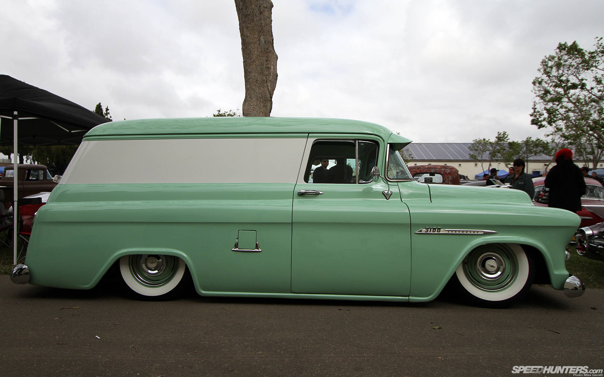 Lowrider 桌布 壁紙 — 一輛薄荷綠色的1950年代復古雪佛蘭廂型車，配備白色輪胎，停放在多雲天空下的戶外車展中。