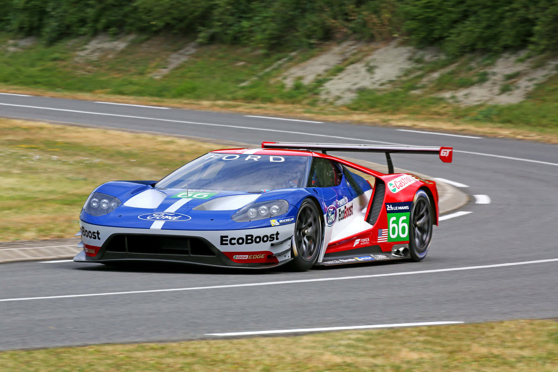 Car Racing 桌布 壁紙 — 一輛藍、紅、白三色的福特GT賽車（編號66）在蜿蜒的瀝青賽道上奔馳，賽道兩旁是綠草和土堤。