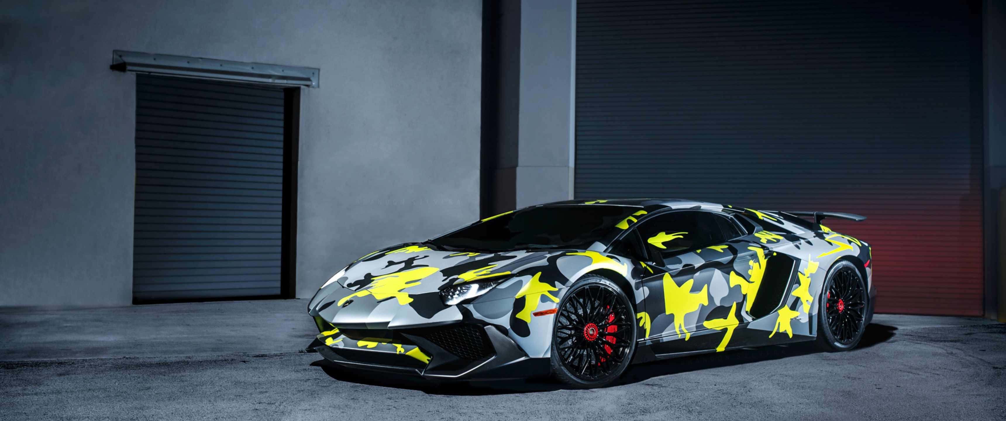 3440x1440p Lamborghini 桌布 壁紙 — 一輛藍寶堅尼 Aventador 帶有醒目的黃色和灰色迷彩塗裝，停在昏暗的工業車庫中。