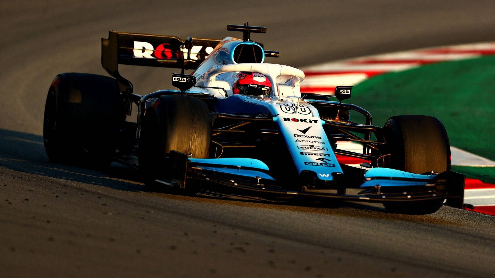 F1 桌布 壁紙 — 一輛藍白相間的F1賽車在陽光普照下，繞過賽道彎角，路肩呈紅、白、綠色。