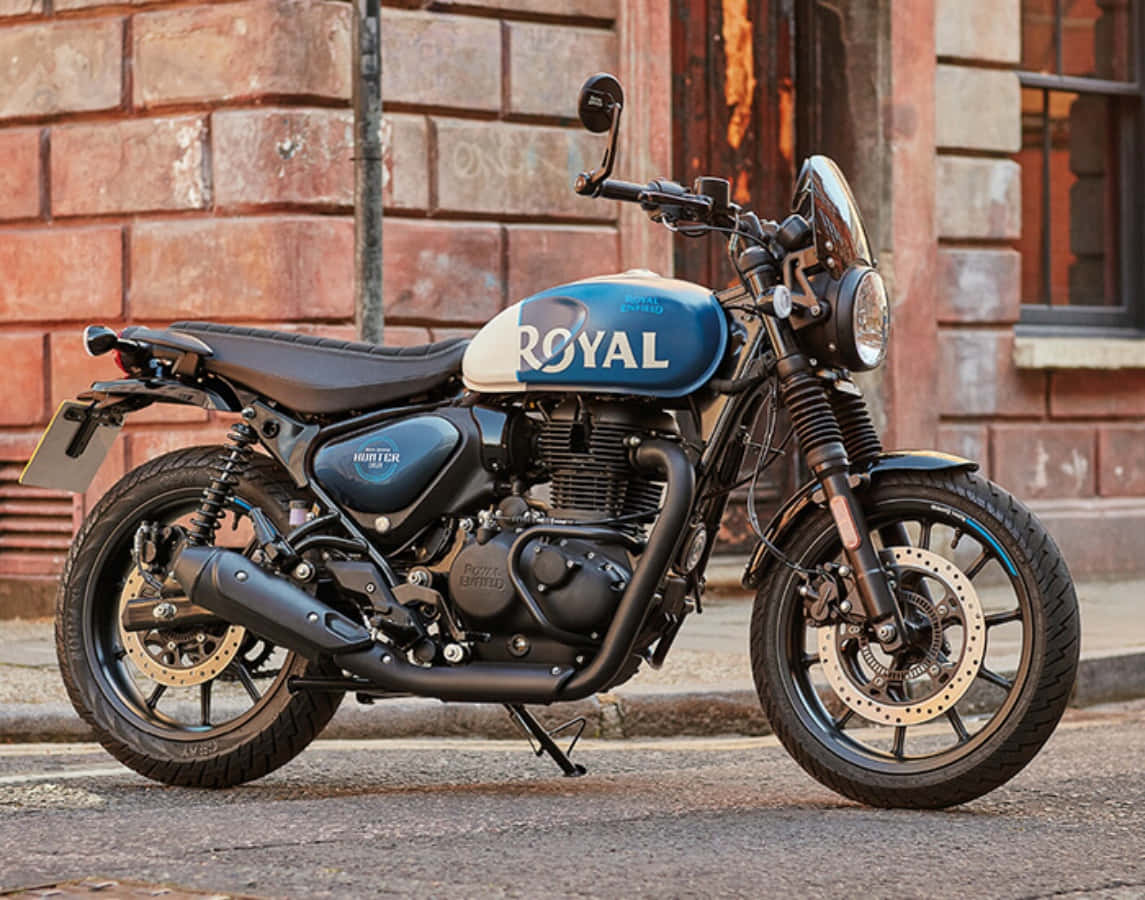 機車 桌布 壁紙 — 一輛藍白色 Royal Enfield Hunter 摩托車停放在鵝卵石街道上，旁邊是一棟風化的磚建築。