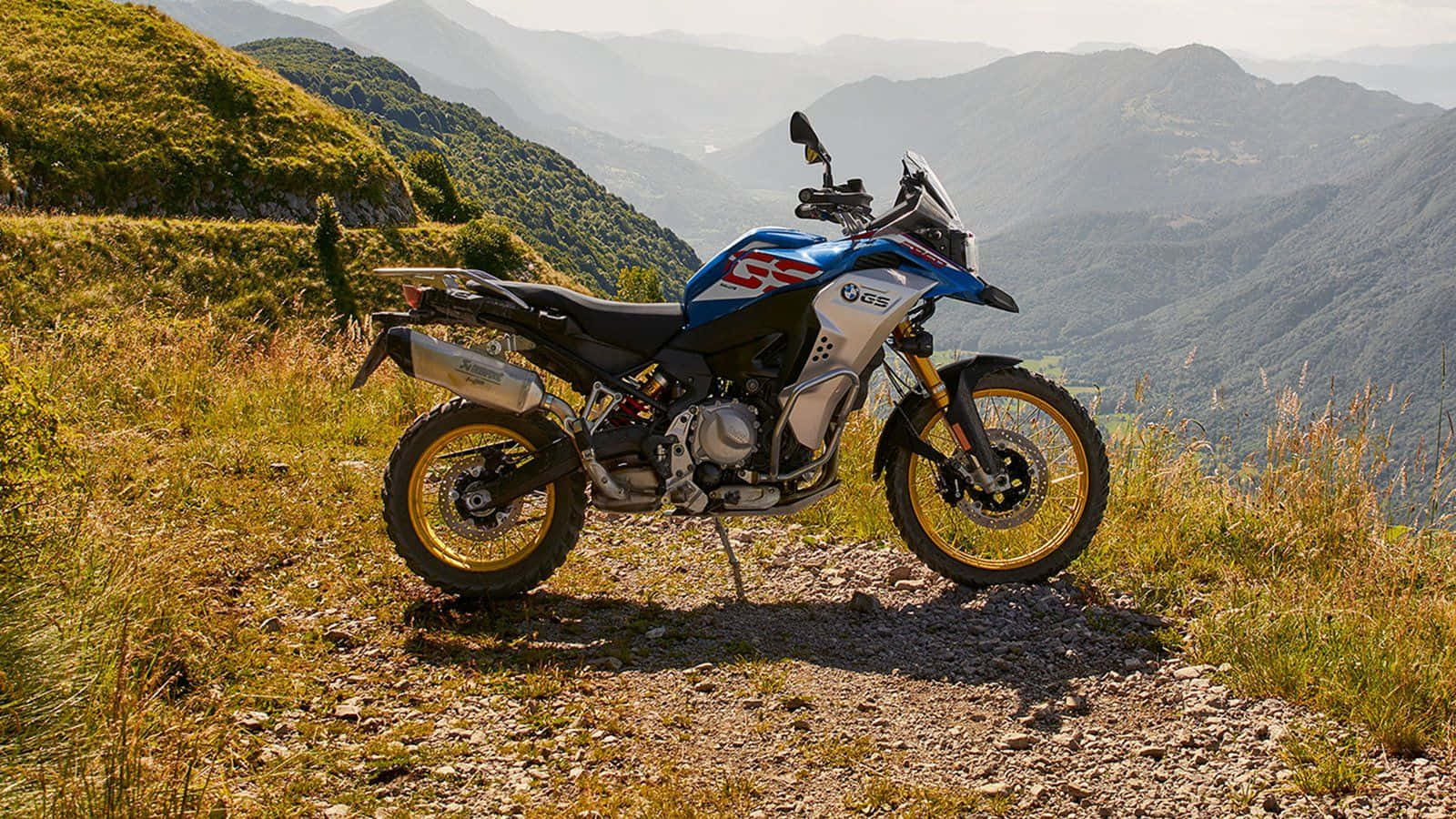 Thrill 桌布 壁紙 — 一輛藍白色的BMW GS冒險摩托車停泊在岩石山徑上，俯瞰著陽光下起伏的綠色山丘。
