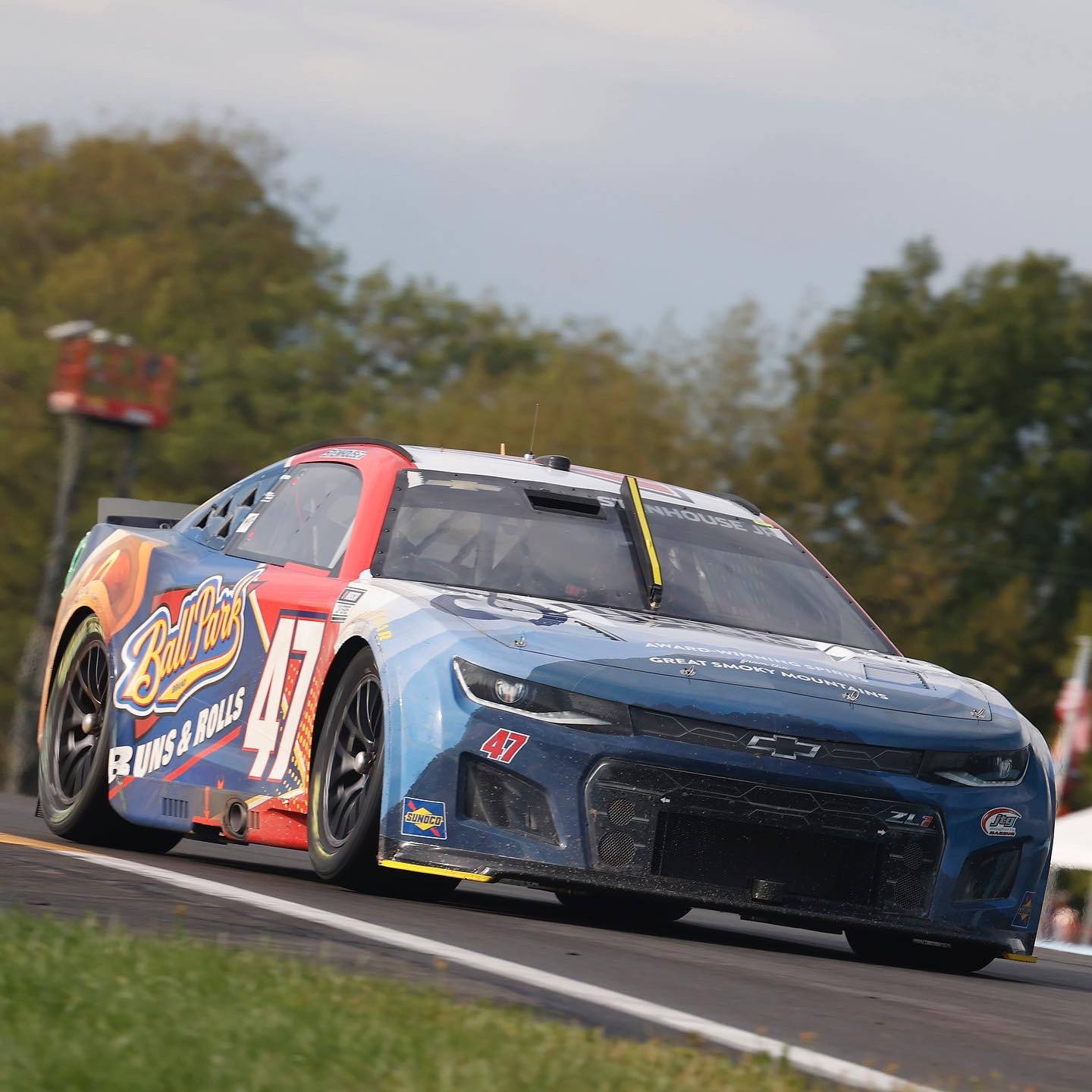 Racing 桌布 壁紙 — 一輛藍紅色的NASCAR賽車（47號）在陰天之下，沿著綠樹成蔭的賽道飛馳。