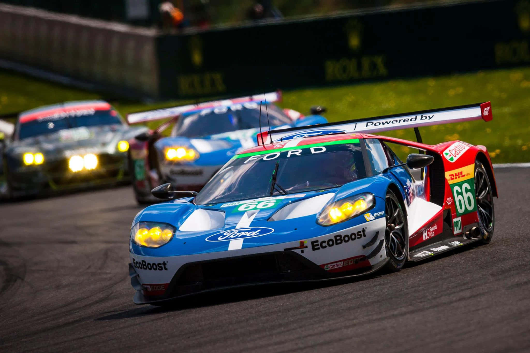 Car Racing 桌布 壁紙 — 一輛藍紅色的福特GT賽車（66號）在賽道上疾馳，車頭燈閃爍，其他賽車緊隨其後。