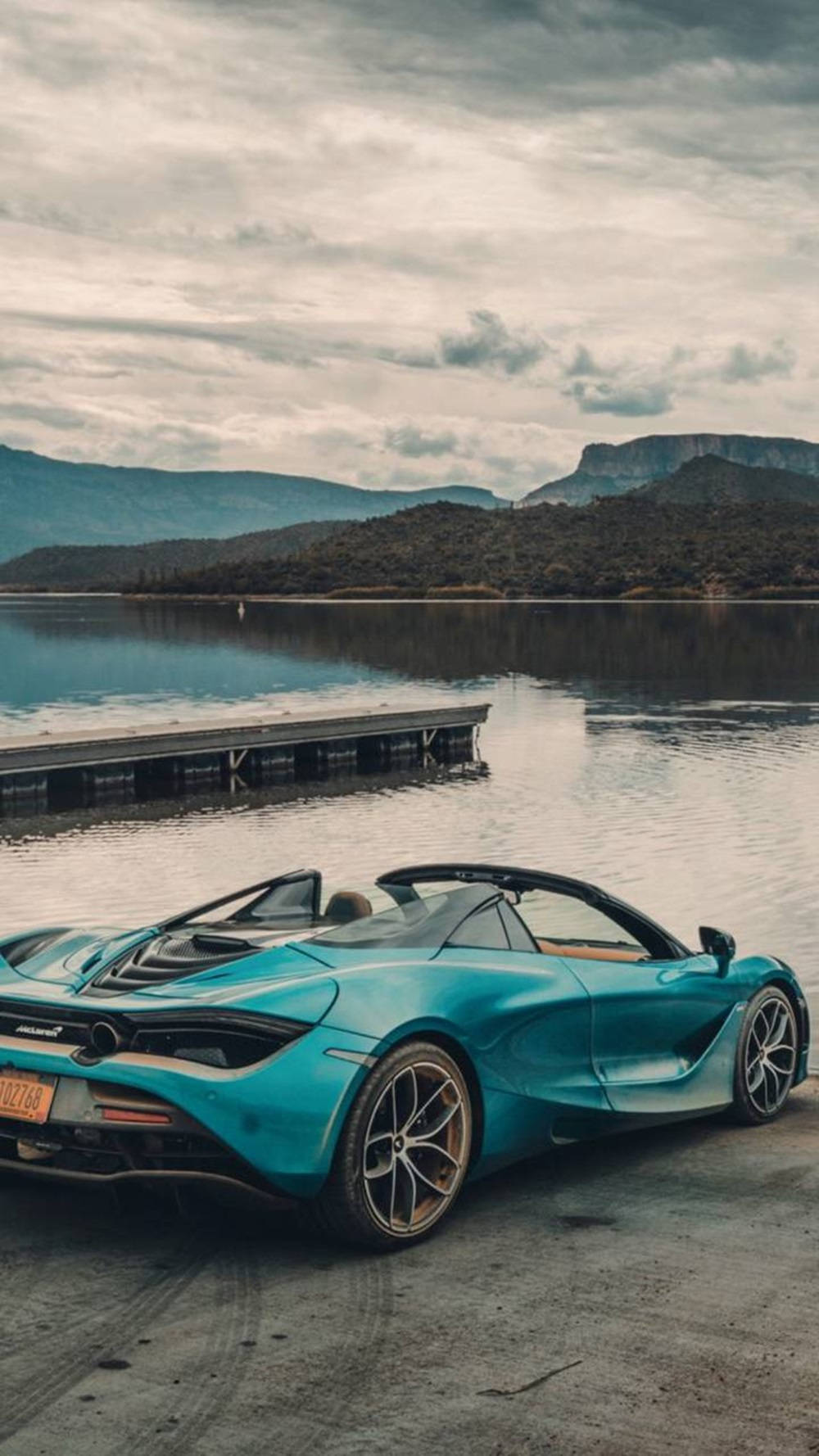 Lake 桌布 壁紙 — 一輛藍綠色McLaren跑車停泊在碼頭旁，旁邊是平靜的湖泊，背景有山脈，天空多雲。