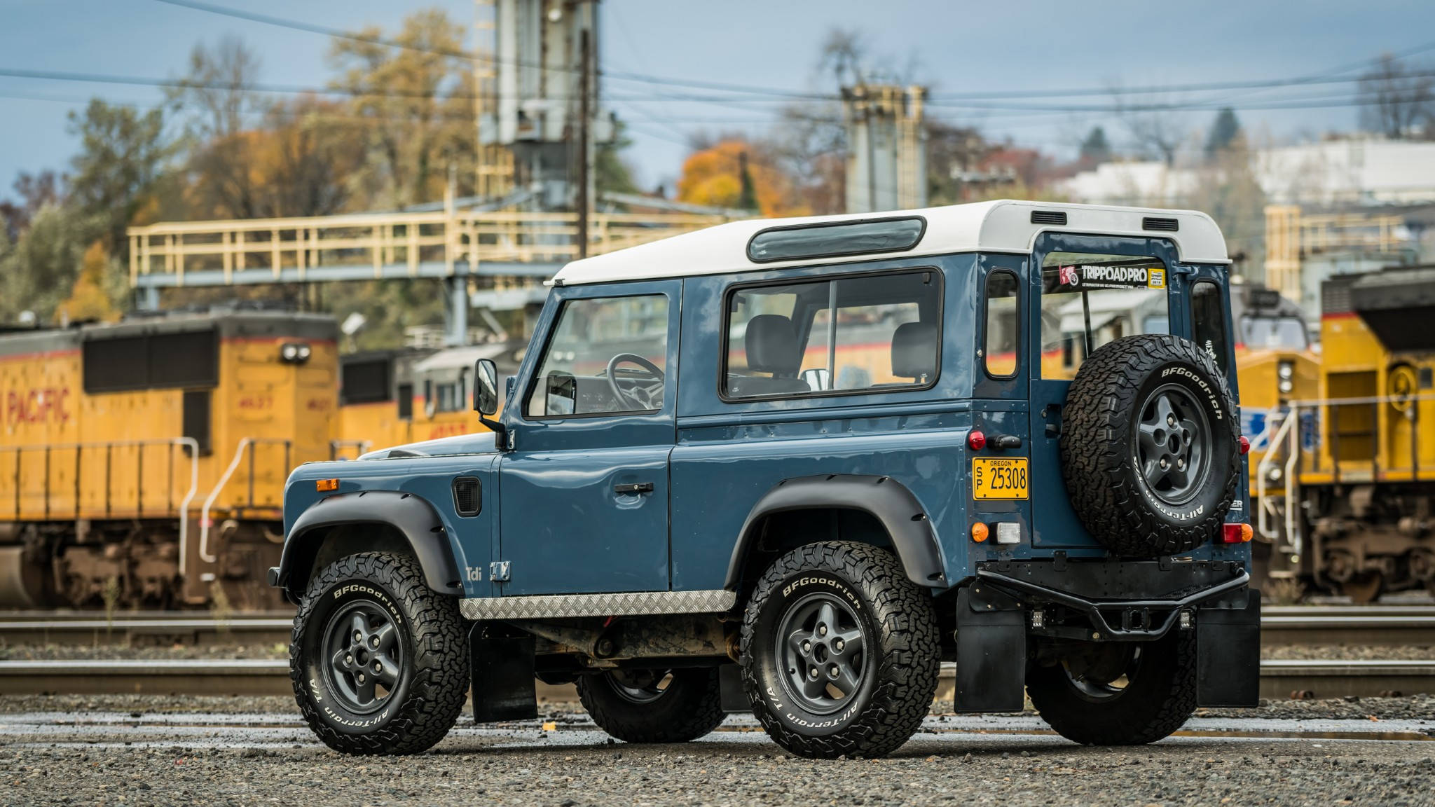 land rover iPhone 桌布 壁紙 — 一輛藍色 Land Rover Defender 堅固地停在鐵軌旁的礫石上，背景有黃色貨運車廂和晴朗的天空。