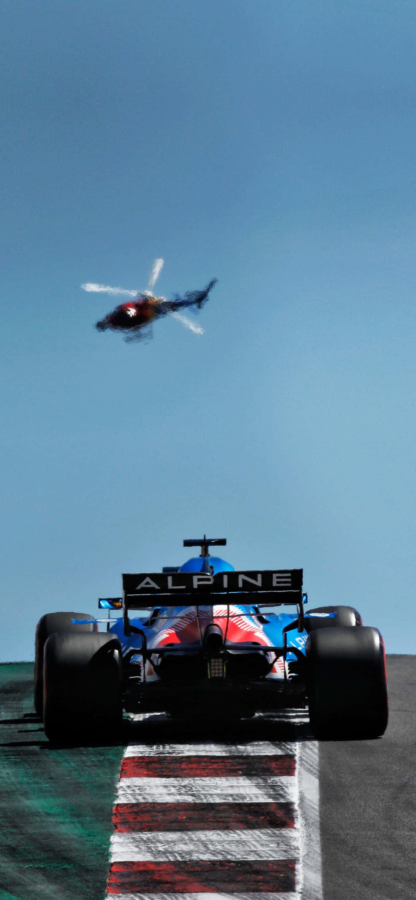 Formula 1 iPhone 桌布 壁紙 — 一輛藍色Alpine一級方程式賽車在賽道上奔馳，一架直升機在晴朗的藍天下飛過。