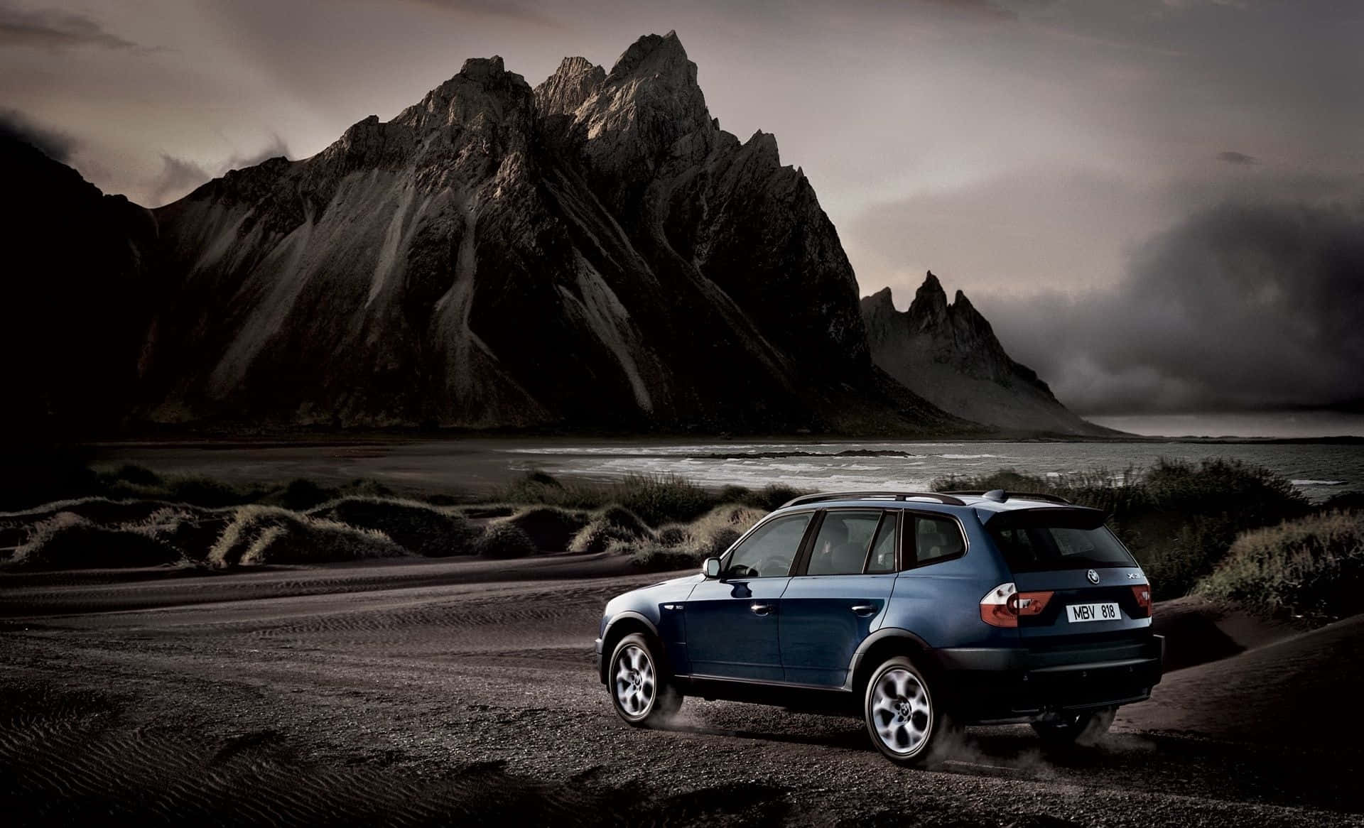 Bmw X3 桌布 壁紙 — 一輛藍色BMW SUV在戲劇性的黑暗山脈和暴風雨天空下行駛在碎石路上。