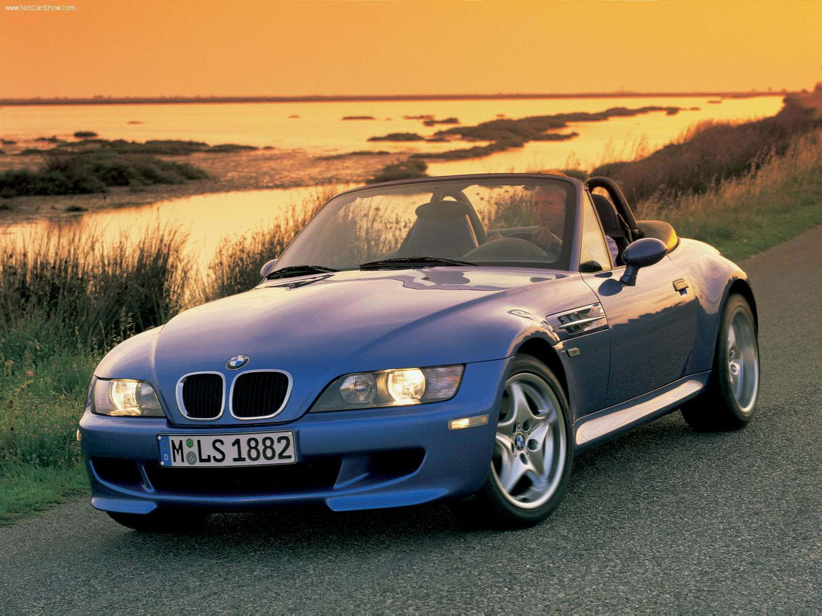 Bmw Z3 桌布 壁紙 — 一輛藍色BMW Z3敞篷跑車在金色日落時分，背景有沼澤地的海岸公路上閃閃發光。