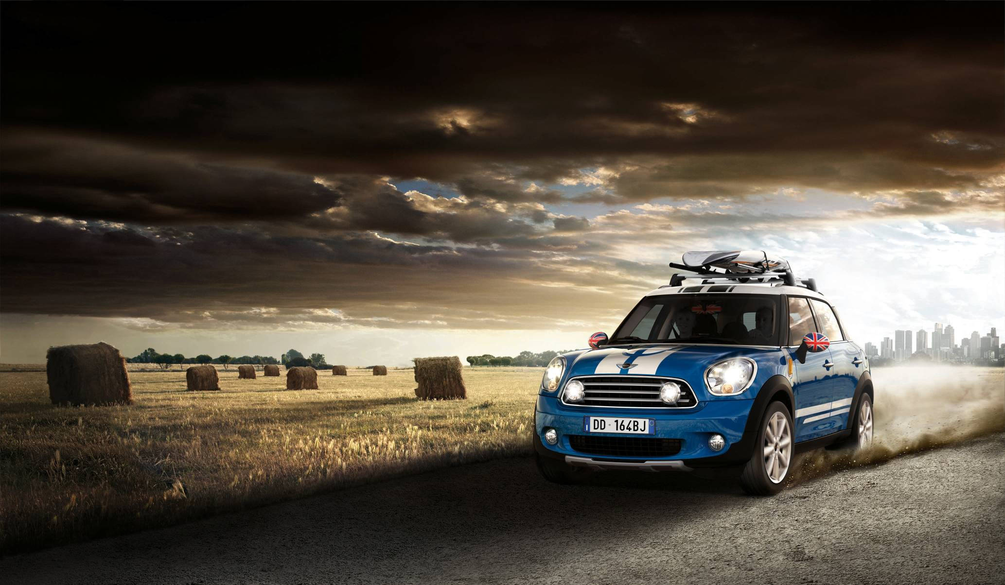 Mini Cooper 桌布 壁紙 — 一輛藍色MINI Countryman在鄉村道路上行駛，揚起灰塵，天空多雲戲劇化，有乾草捆和遠處的城市天際線。