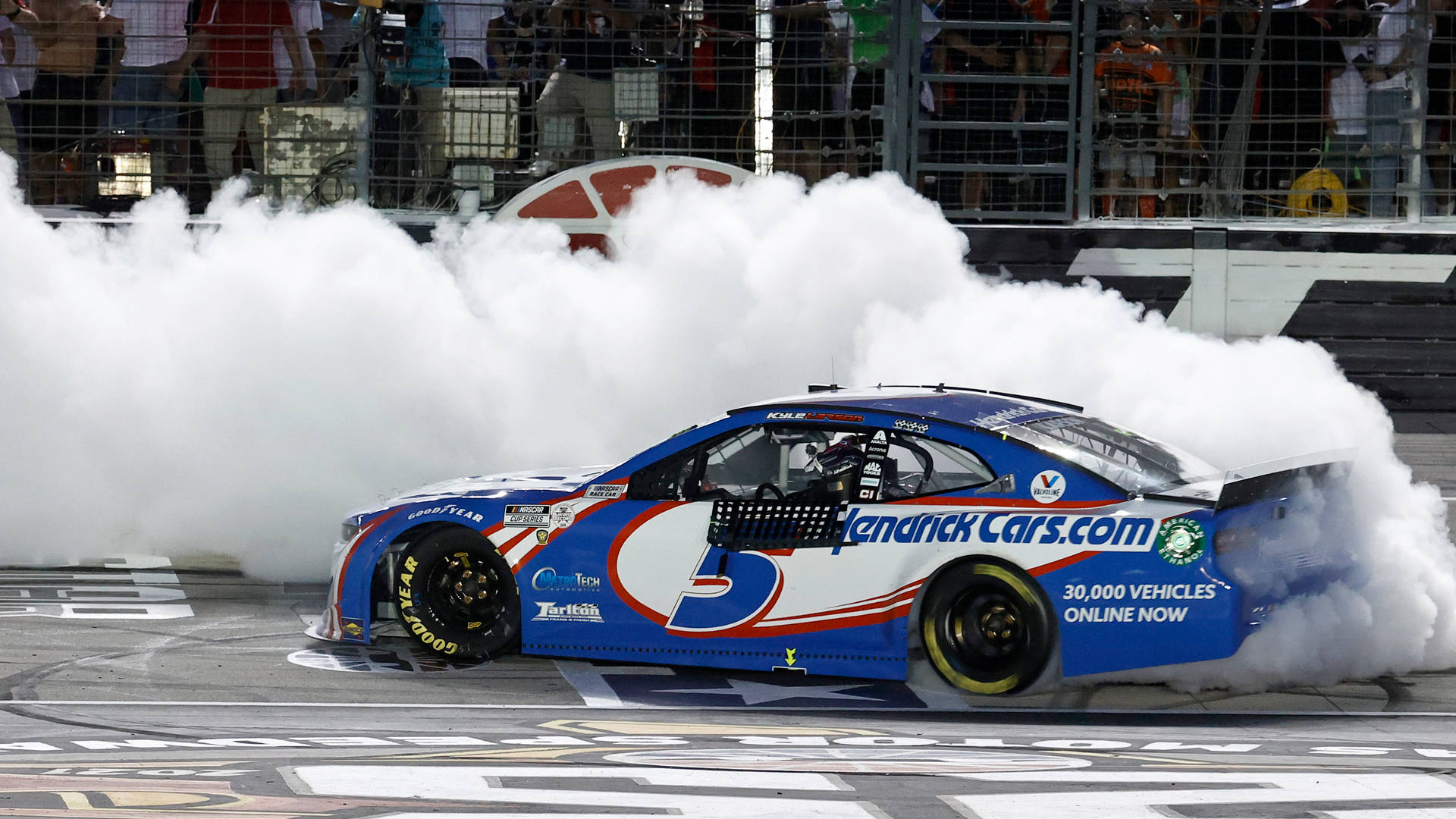 Kyle Larson 桌布 壁紙 — 一輛藍色NASCAR賽車在明亮的體育場燈光下戲劇性地冒煙衝過終點線。