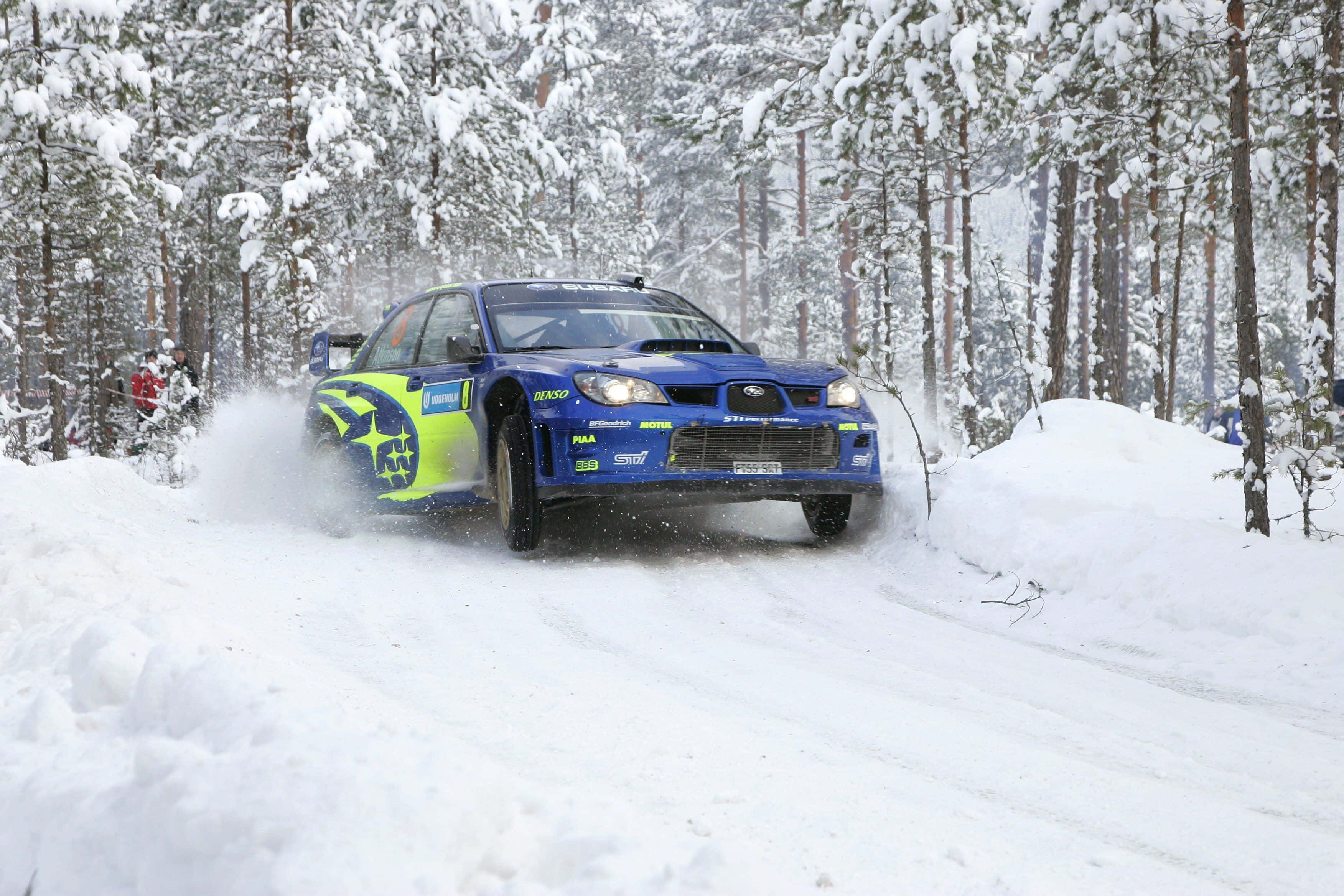 Ice Racing 桌布 壁紙 — 一輛藍色和螢光綠色的拉力賽車在雪地森林賽道上疾馳，揚起雪霧。