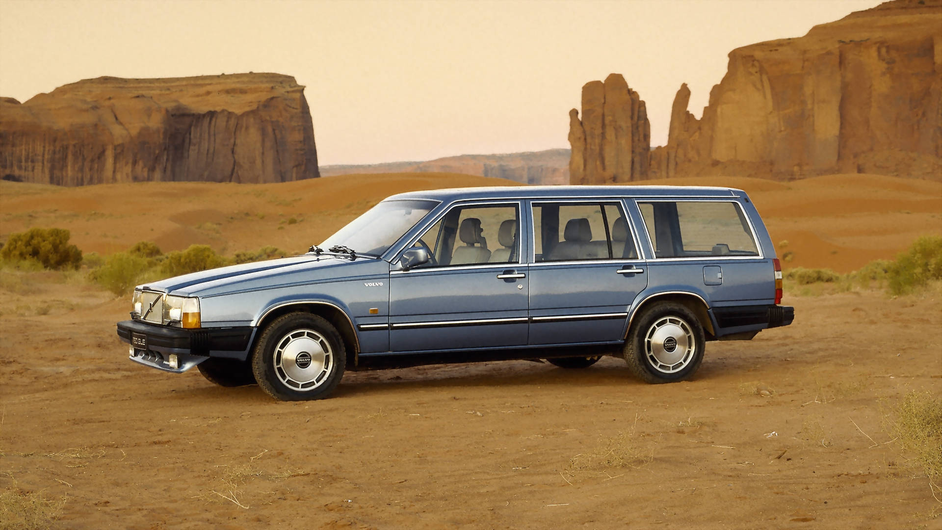 volvo iPhone 桌布 壁紙 — 一輛藍色富豪旅行車停在佈滿灰塵的地形上，背景是朦朧天空下高聳的紅色岩層。