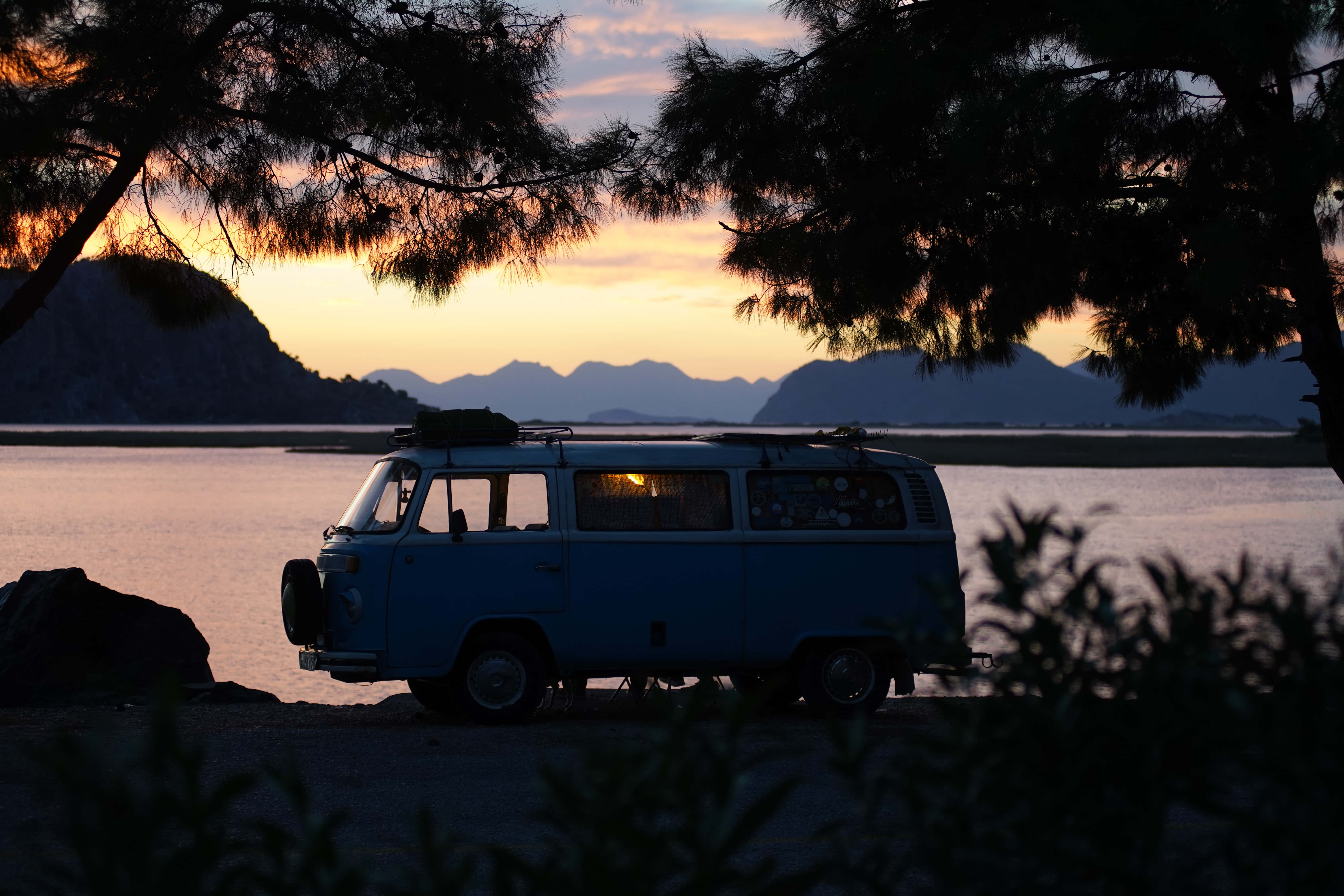 road trip 桌布 壁紙 — 一輛藍色復古廂型車停在沙灘上，夕陽西下，背景是群山，被樹枝框住。