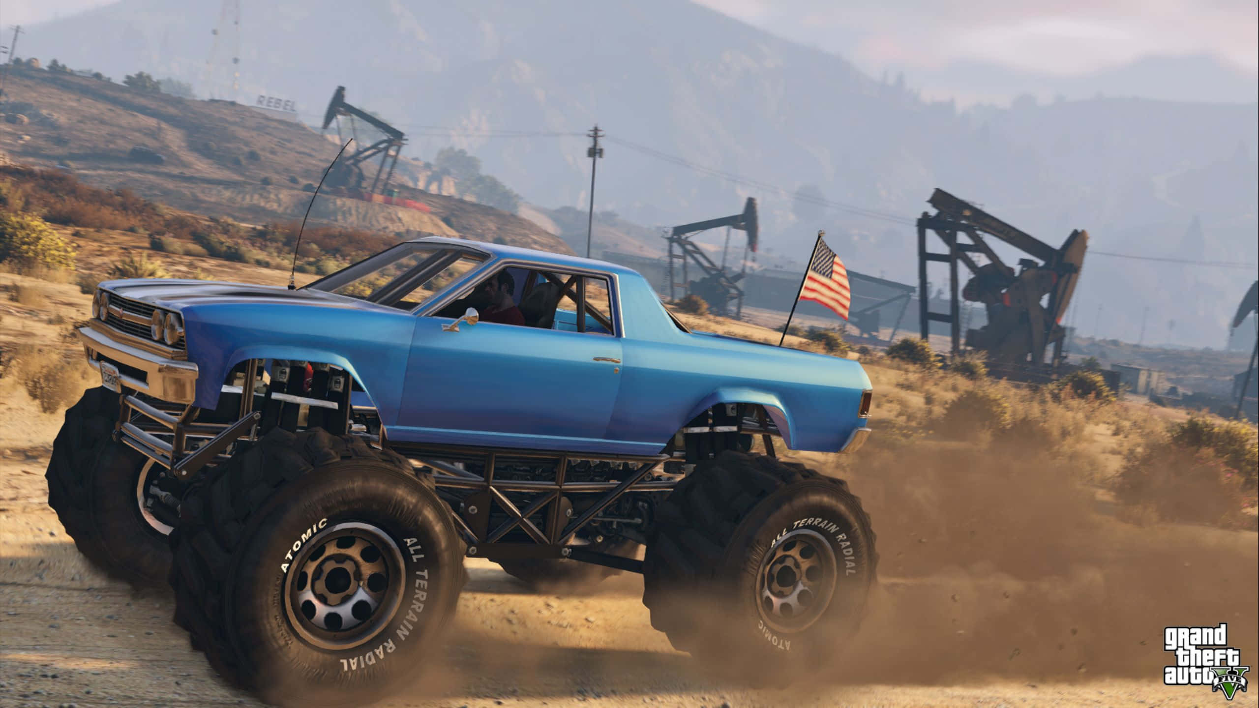 Grand Theft Auto V 桌布 壁紙 — 一輛藍色怪獸卡車配備巨型輪胎，在塵土飛揚的沙漠景觀中咆哮，背景有油泵。