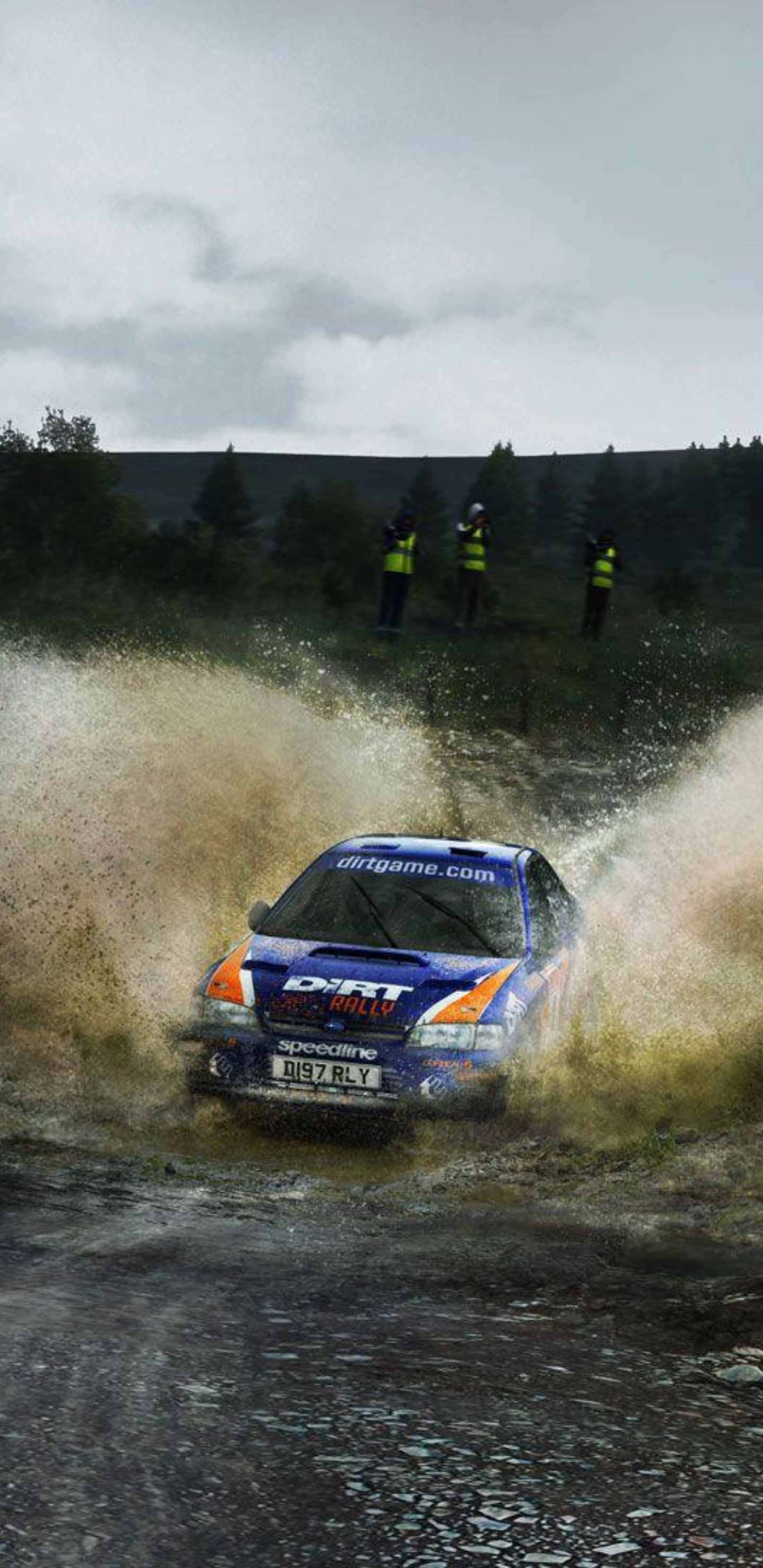 Android Dirt Rally 桌布 壁紙 — 一輛藍色拉力賽車在泥濘的林間小徑上飛馳，在多雲的天空下濺起棕色的水和泥土。