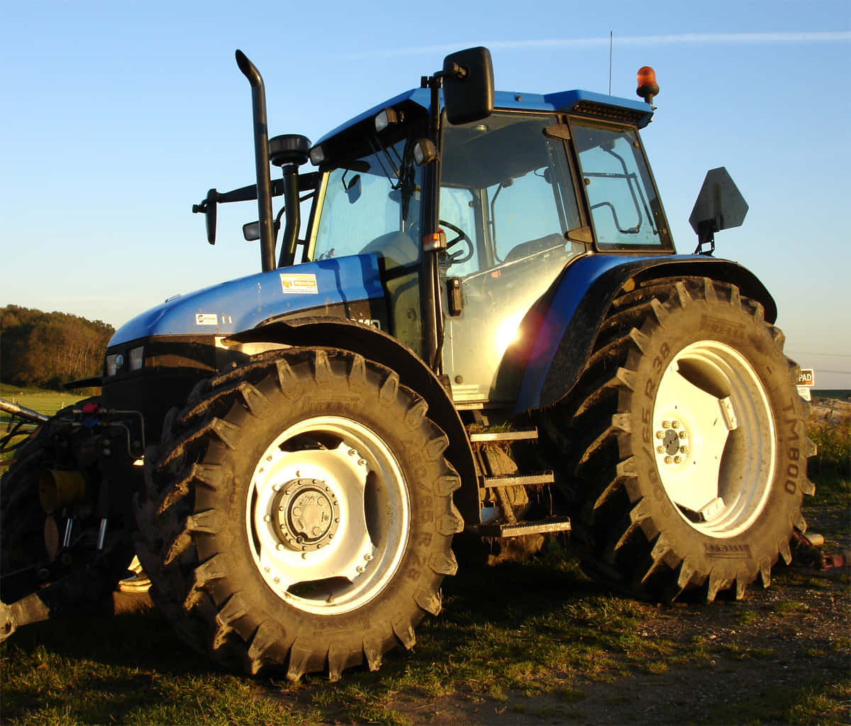 Tractor 桌布 壁紙 — 一輛藍色拖拉機配備巨大胎紋輪胎，在黃金時段晴朗天空下的草地上。