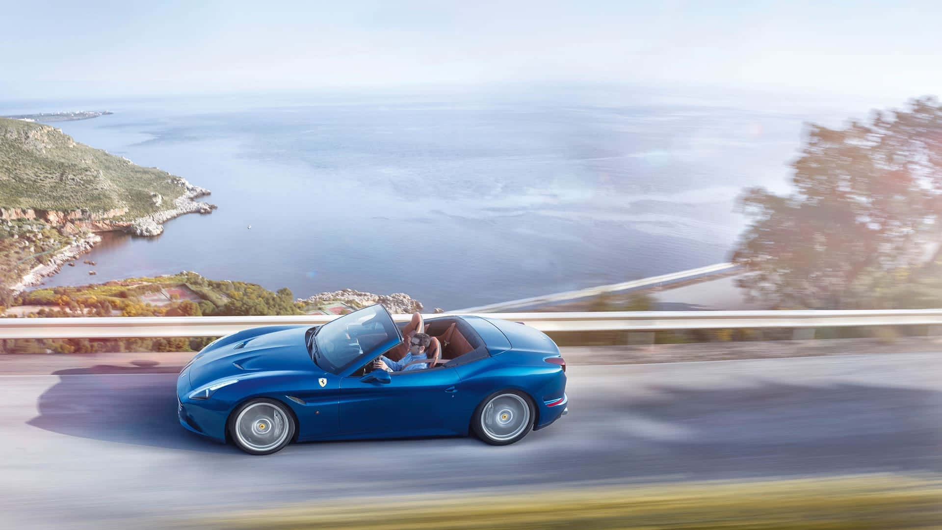 Ferrari California T 桌布 壁紙 — 一輛藍色法拉利敞篷車沿著海岸公路疾馳，司機揮手致意，俯瞰廣闊的海洋和陽光普照的懸崖。
