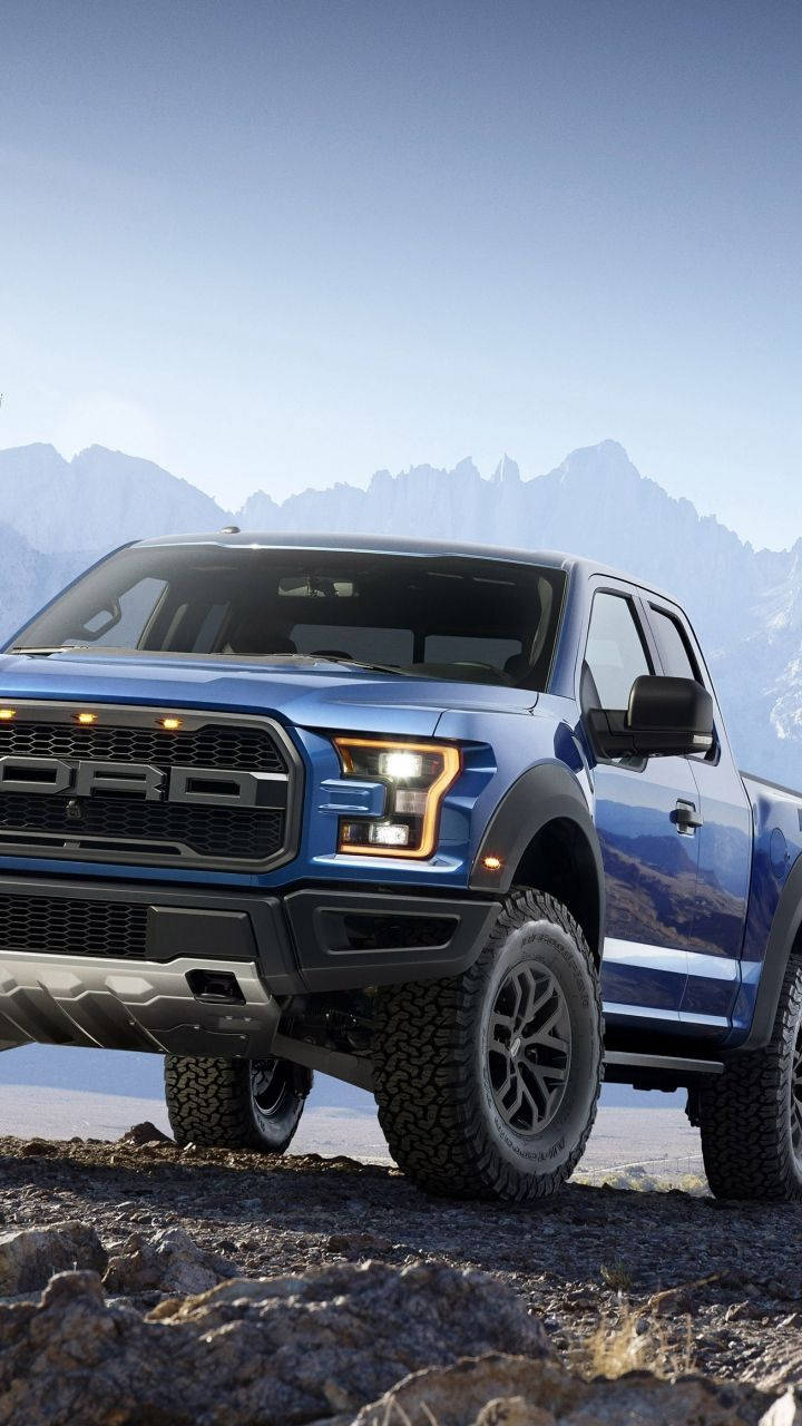 Trucks 桌布 壁紙 — 一輛藍色福特 F-150 Raptor 卡車，車頭燈發光，停在岩石地形上，背景是晴朗天空下遠處的雪山。