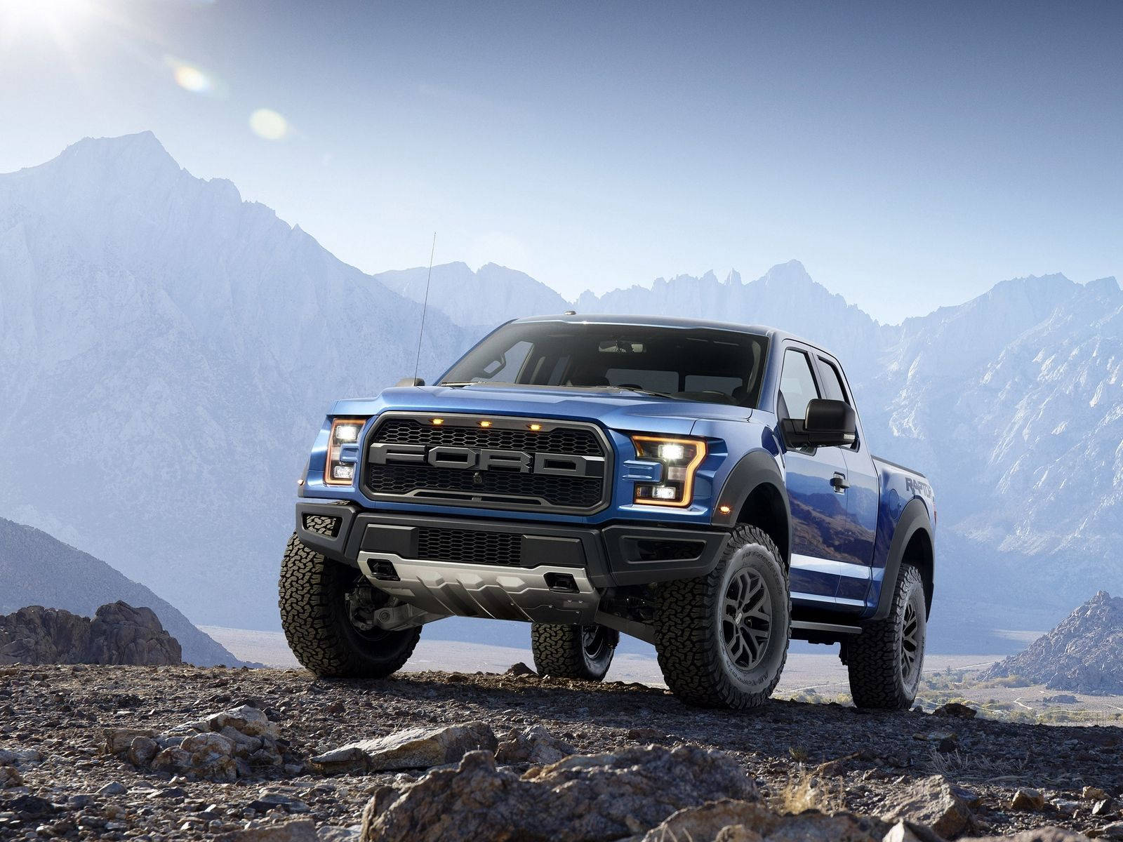 Raptor 桌布 壁紙 — 一輛藍色福特F-150 Raptor卡車停在岩石地形上，背後有白雪覆蓋的山脈在明亮的陽光下聳立。