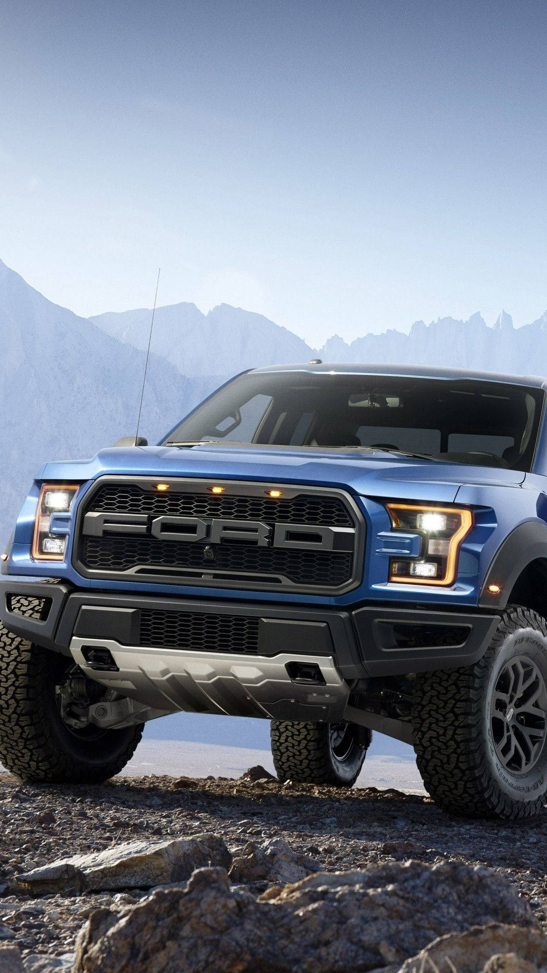 ford iPhone 桌布 壁紙 — 一輛藍色福特F-150猛禽卡車，車頭燈發光，站在岩石地形上，背景是遠山和晴朗的天空。