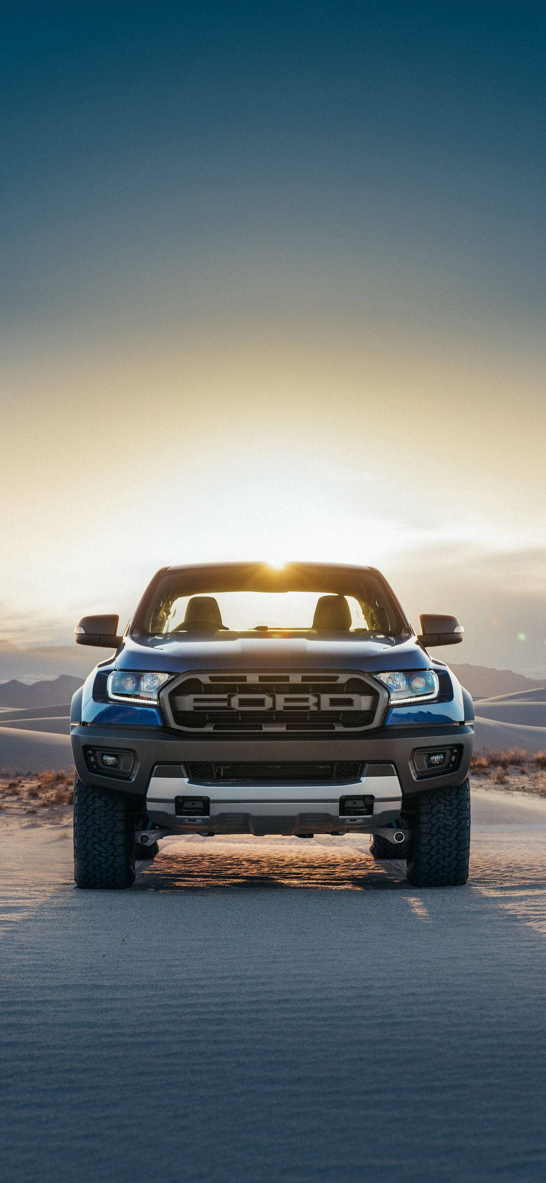 ford iPhone 桌布 壁紙 — 一輛藍色福特卡車在沙漠公路上向前行駛，夕陽西下，車頭燈閃爍，遠山連綿。