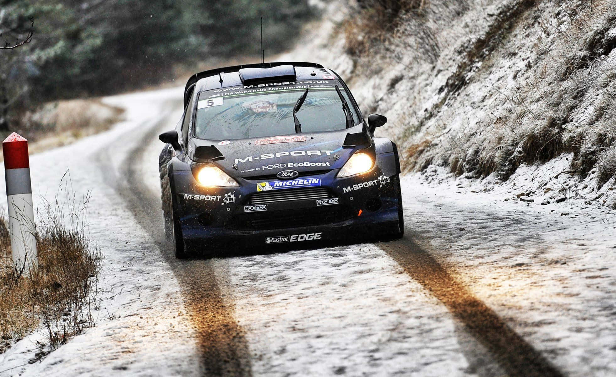 Ice Racing 桌布 壁紙 — 一輛藍色福特拉力賽車亮著頭燈，在白雪覆蓋的森林道路上飛馳，兩旁是積雪的懸崖和一根紅白相間的柱子。