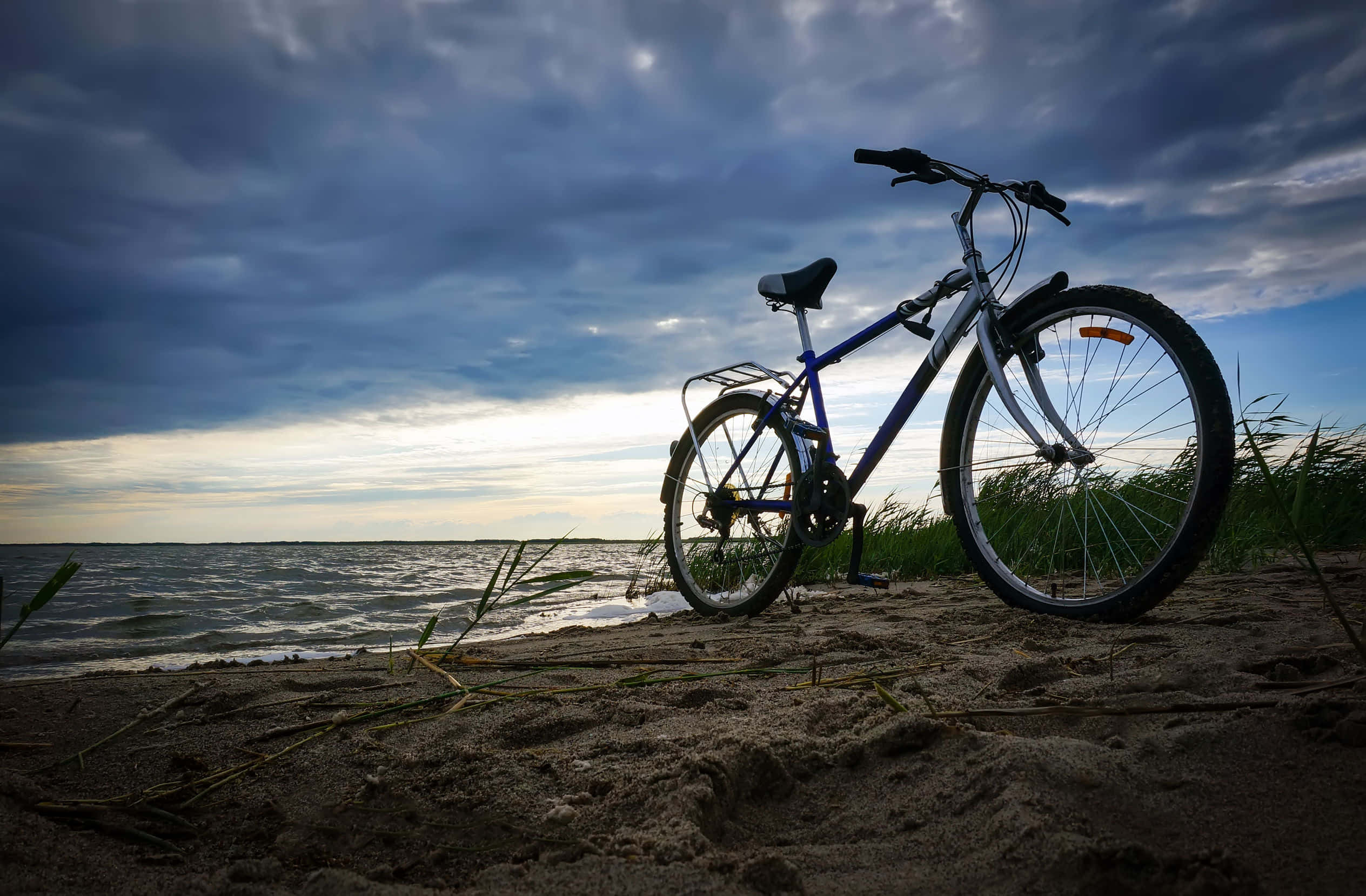 Bicycle 桌布 壁紙 — 一輛藍色腳踏車在黃昏時的沙灘上呈現剪影，海浪在戲劇性的多雲天空下拍打。