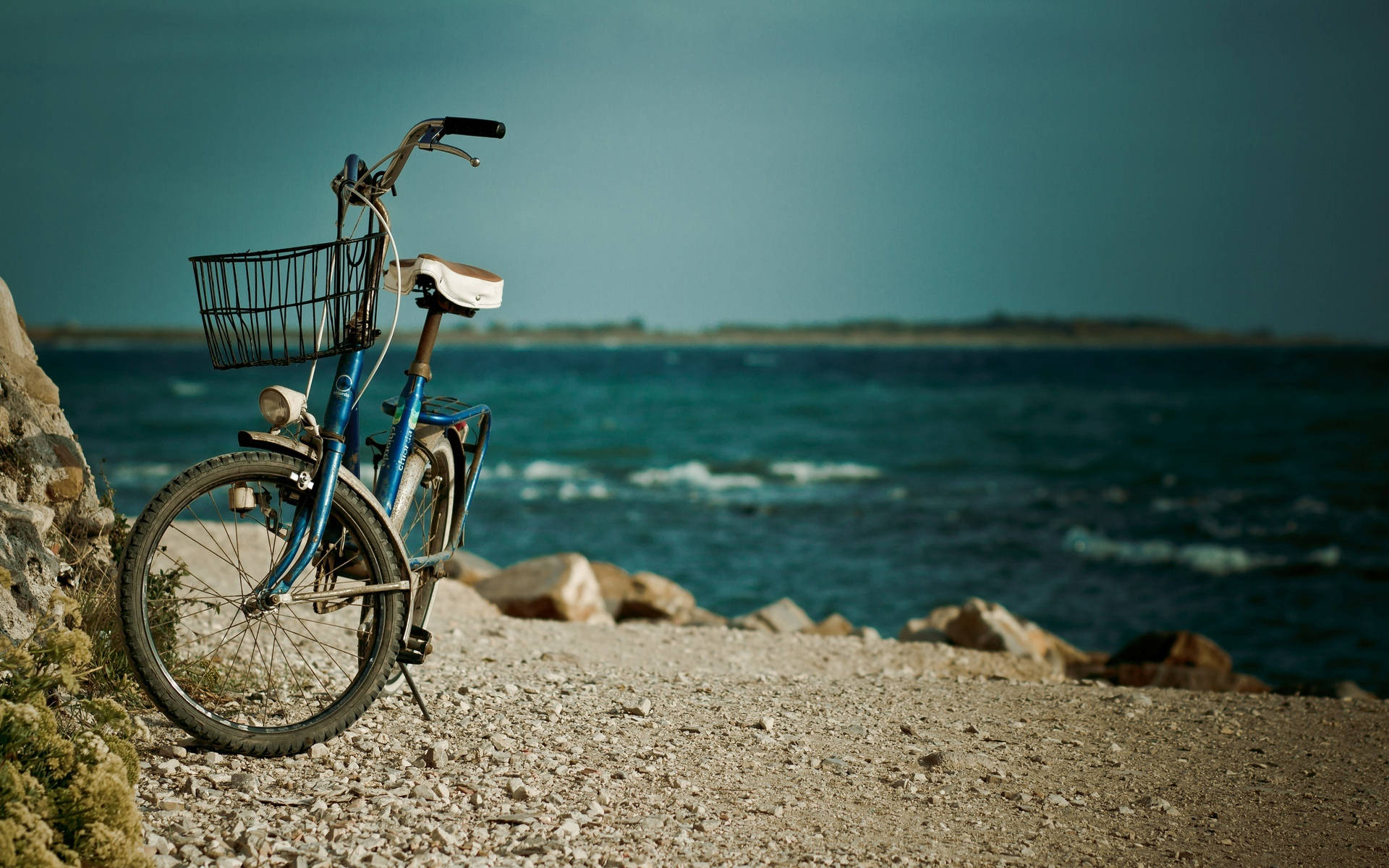 Bicycle 桌布 壁紙 — 一輛藍色自行車帶著籃子靠在石灘的岩石上，俯瞰著暴風雨天空下洶湧的藍綠色大海。
