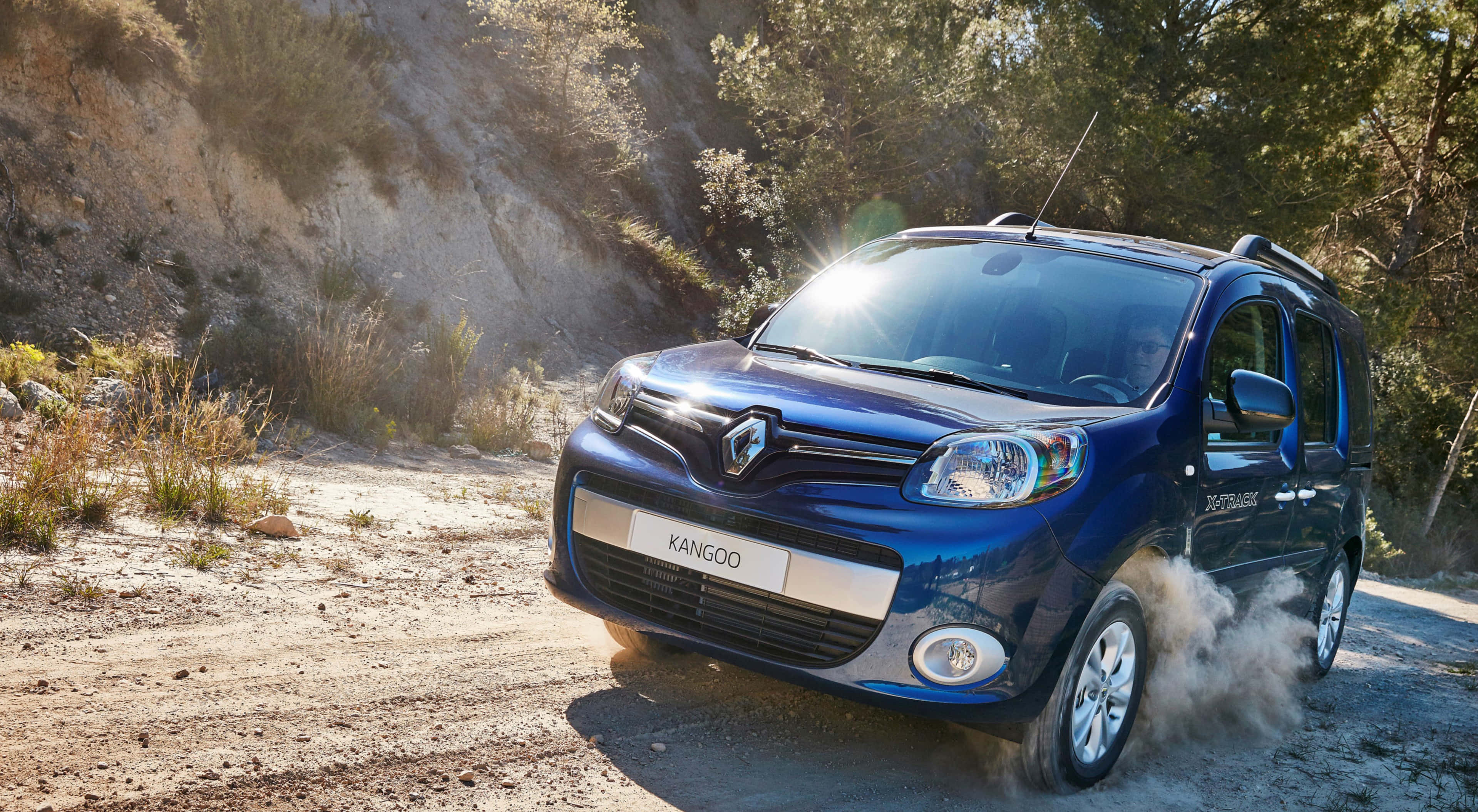 Renault Kangoo 桌布 壁紙 — 一輛藍色雷諾Kangoo在塵土飛揚的地形上行駛，陽光從引擎蓋上反射出來，崎嶇的道路兩旁樹木林立。
