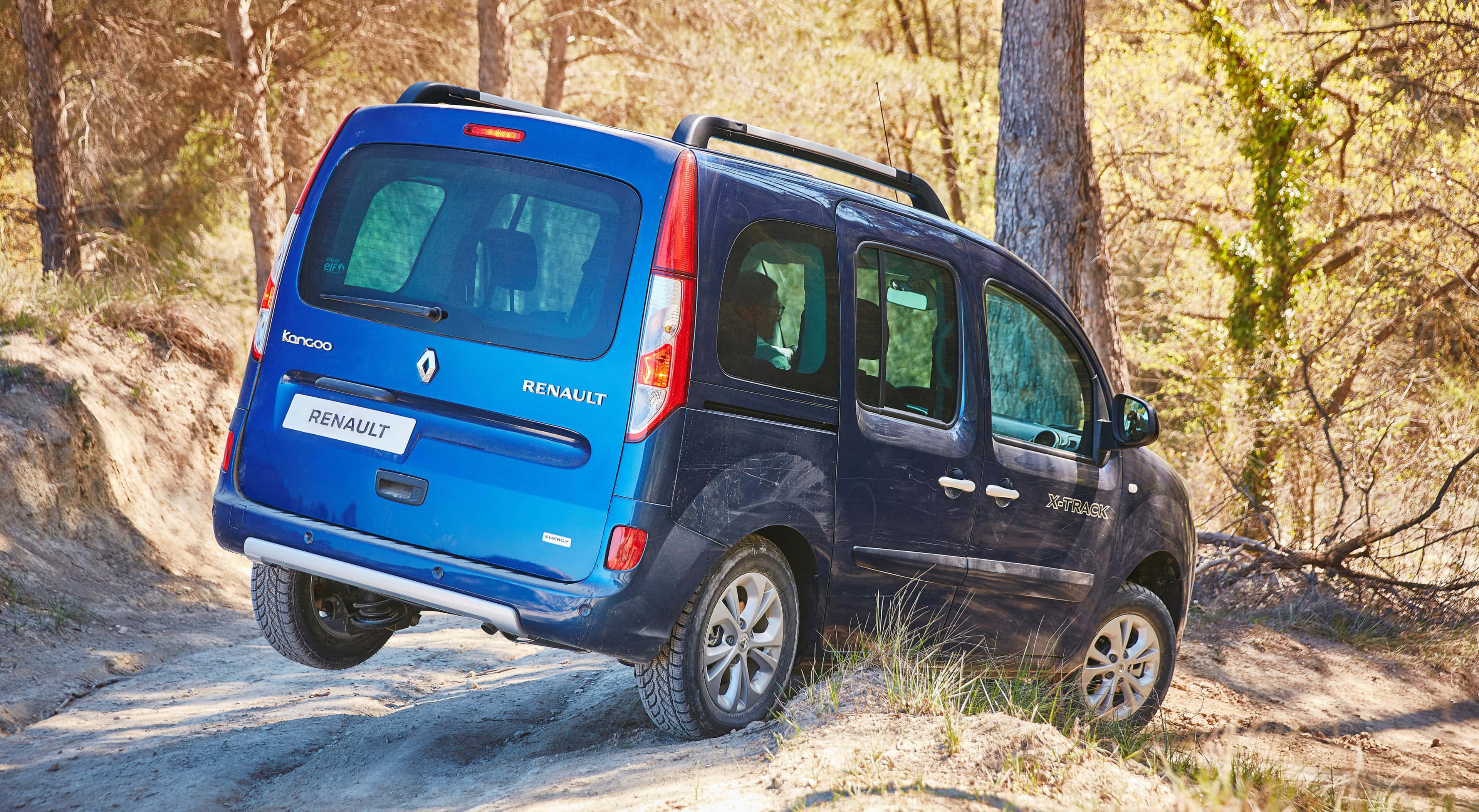 Renault Kangoo 桌布 壁紙 — 一輛藍色雷諾Kangoo廂型車沿著陽光明媚的泥土路穿過高聳樹木和斑駁光影的森林向上行駛。