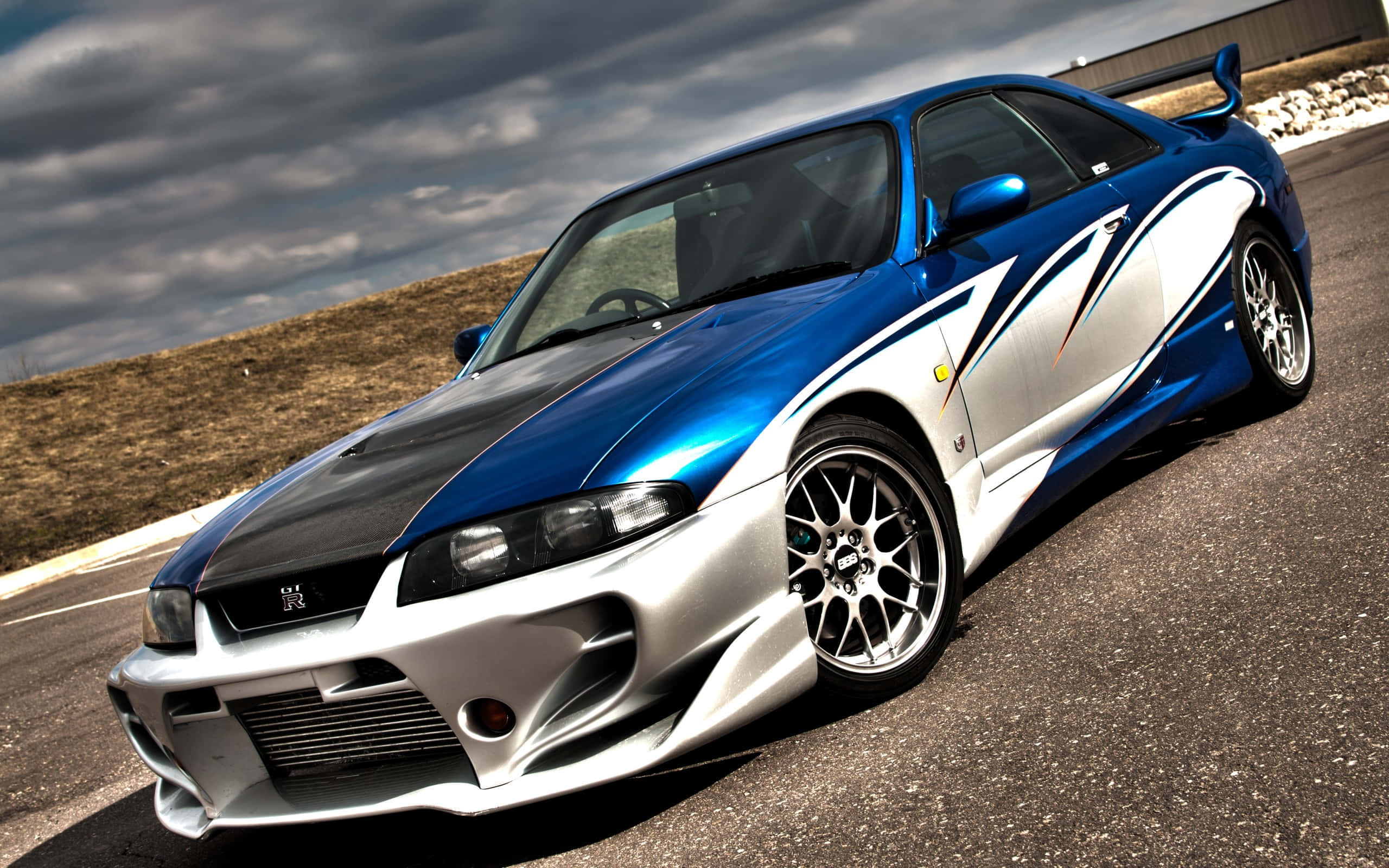R33 GT-R 桌布 壁紙 — 一輛藍銀色日產R34 GT-R跑車帶有白色賽車條紋，停在多雲天空下的瀝青路上。