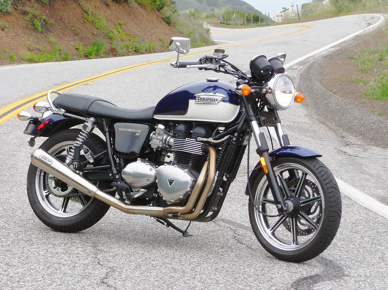 Triumph 桌布 壁紙 — 一輛藍銀色的Triumph摩托車停放在蜿蜒的柏油路上，背景有山丘和稀疏的綠植。