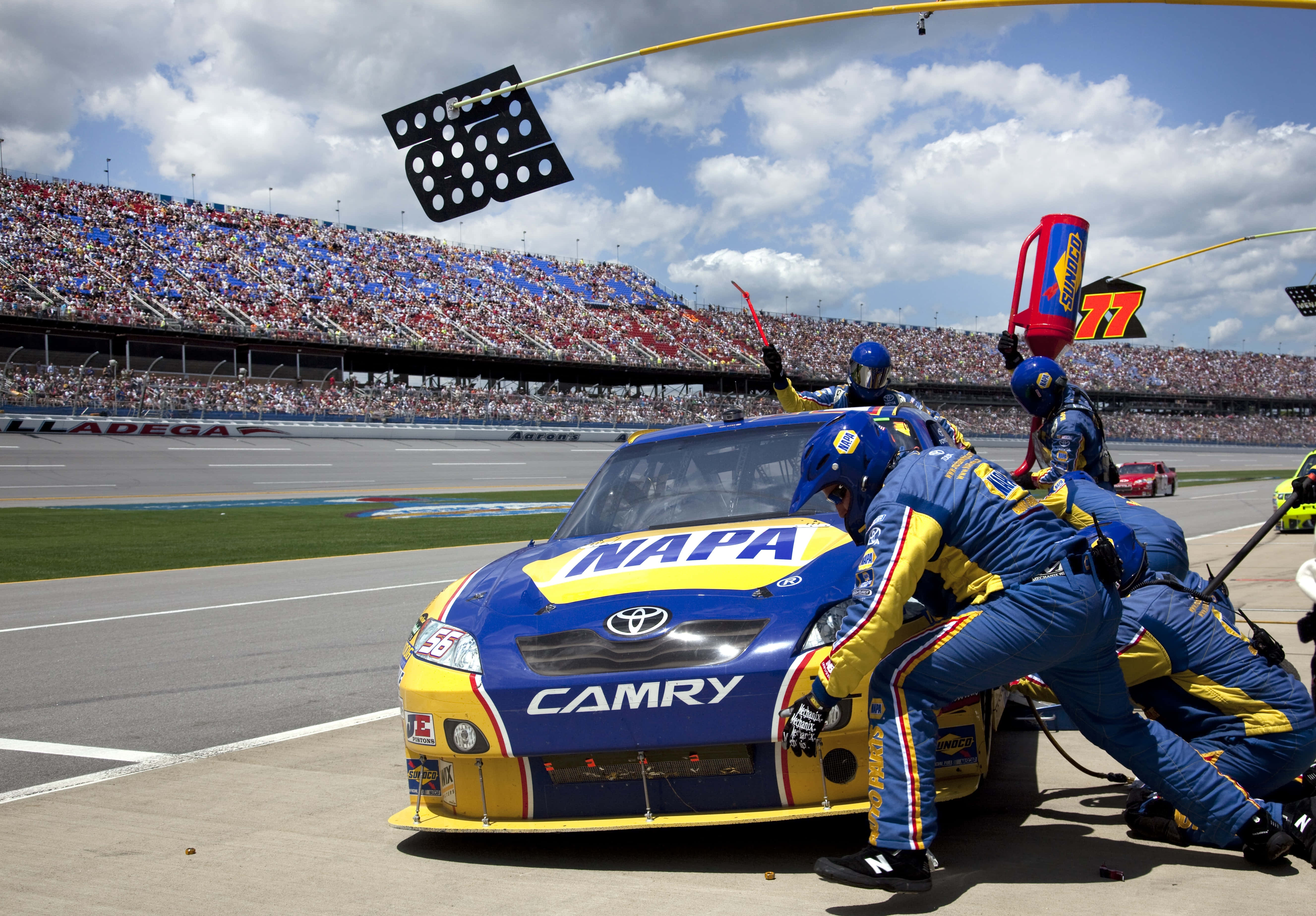 nascar 桌布 壁紙 — 一輛藍黃色的NASCAR豐田Camry賽車正在維修站停靠，身穿藍黃色制服的工作人員在多雲的天空下，於擠滿觀眾的賽道上更換輪胎。