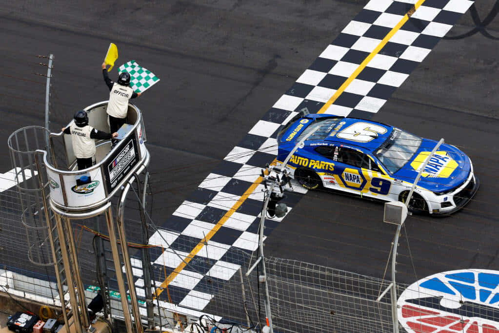 nascar 桌布 壁紙 — 一輛藍黃色的NASCAR賽車越過終點線，工作人員在塔上揮舞旗幟。