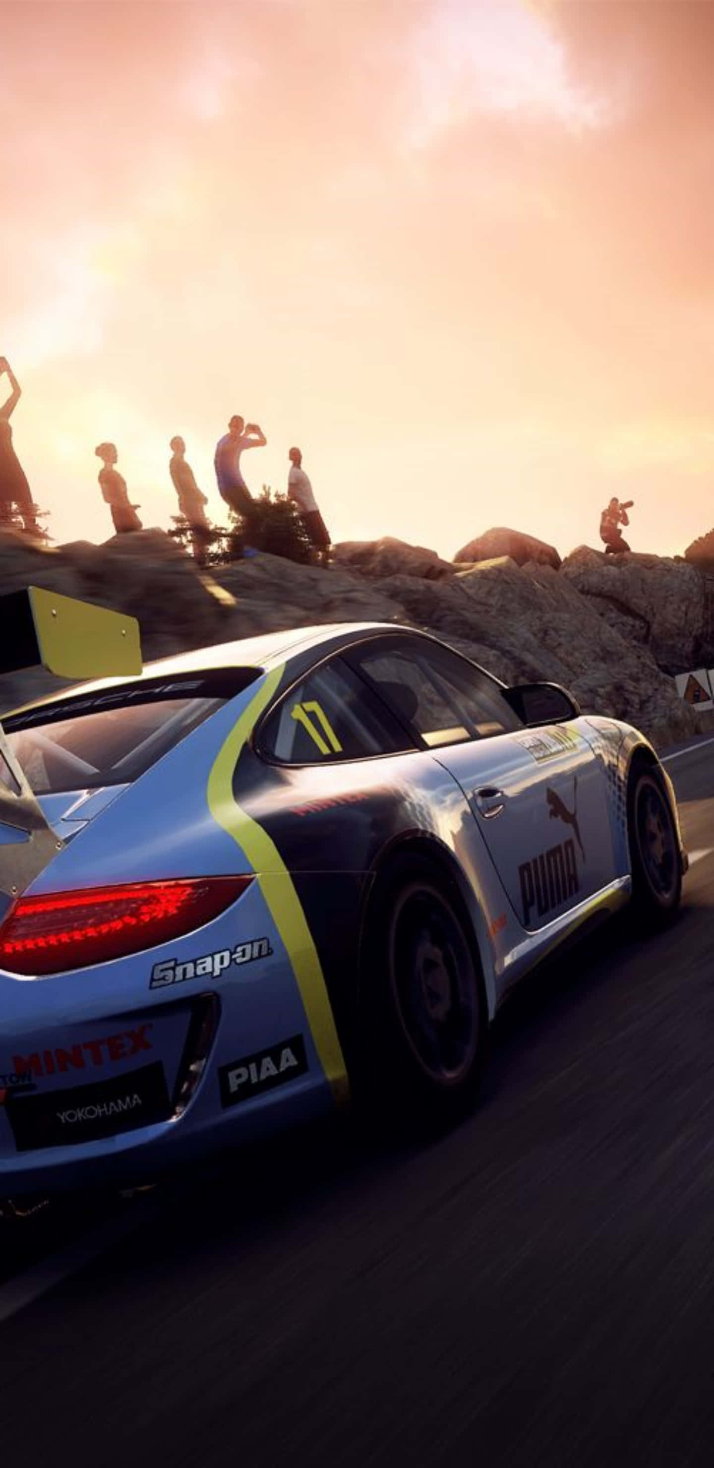 Pixel 3xl Dirt Rally 桌布 壁紙 — 一輛藍黃色賽車在蜿蜒的山路上奔馳，觀眾在日落時分從岩石懸崖上觀看。