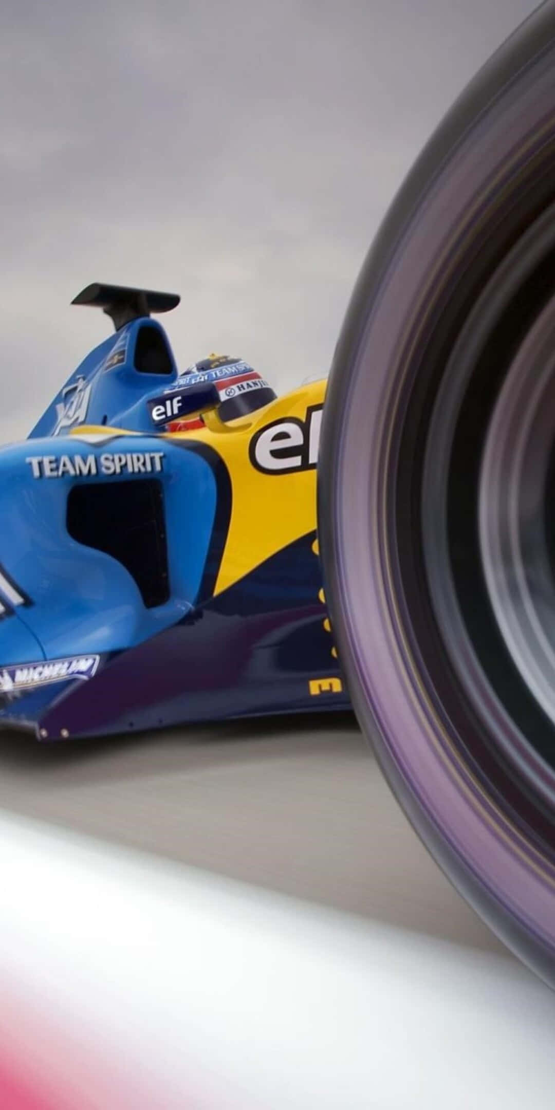 F1 桌布 壁紙 — 一輛藍黃色賽車高速駛過，帶有動態模糊效果，輪胎佔據前景主導位置。