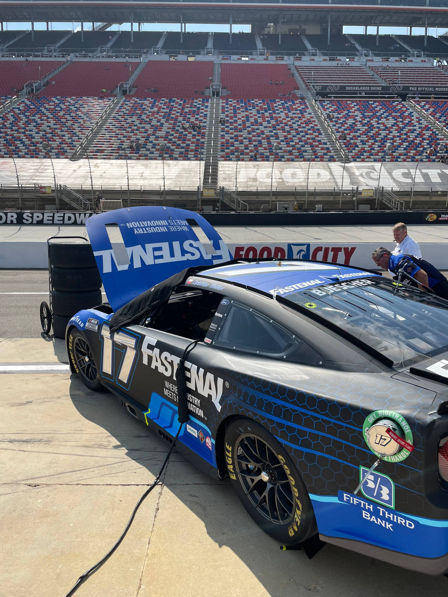 Ford Mustang 桌布 壁紙 — 一輛藍黑相間的NASCAR賽車打開引擎蓋，停在體育場的維修區，在明亮的日光下。