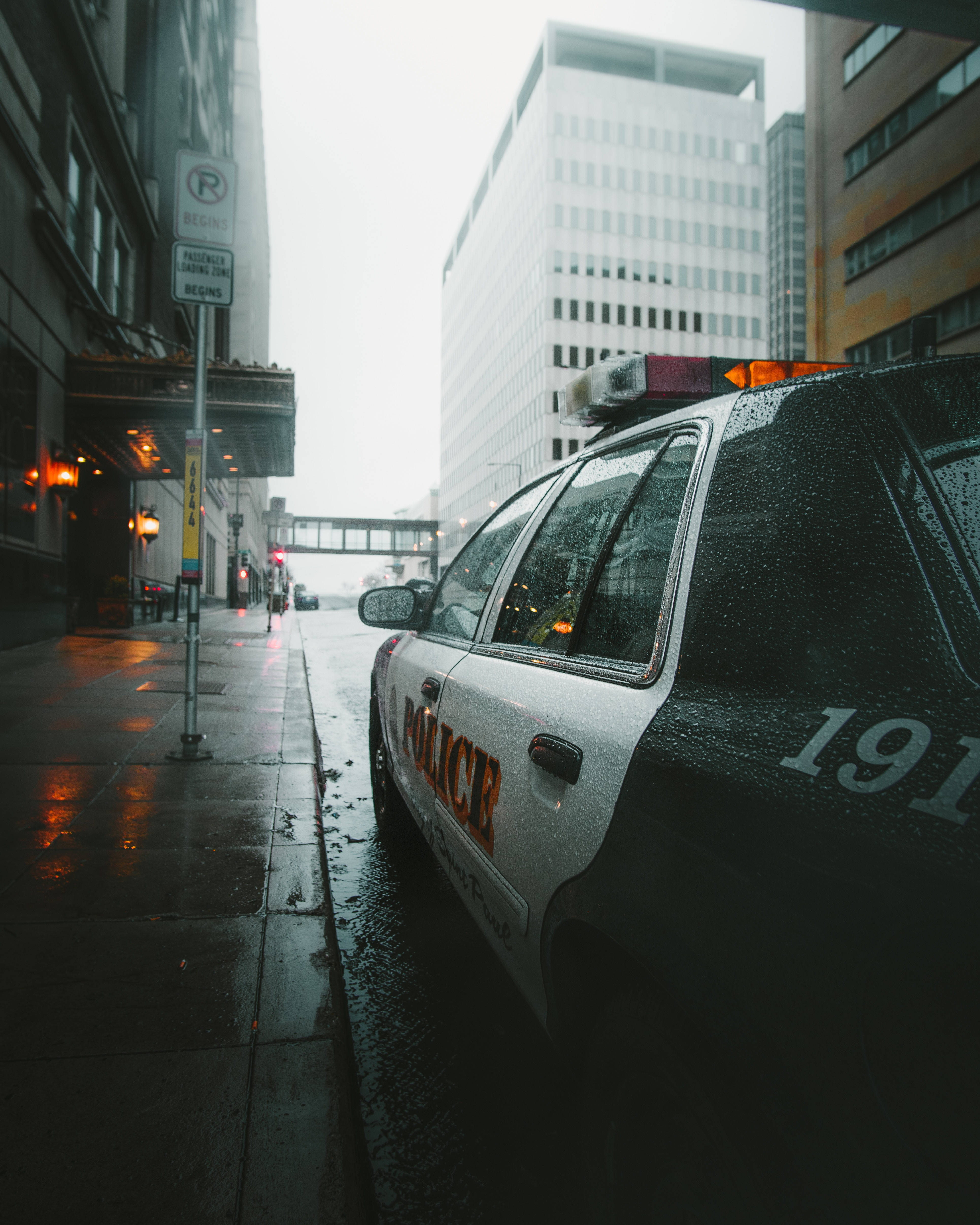 警察 桌布 壁紙 — 一輛被雨水淋濕的警車停泊在濕漉漉的城市街道上，高樓大廈在灰濛濛的天空下聳立。