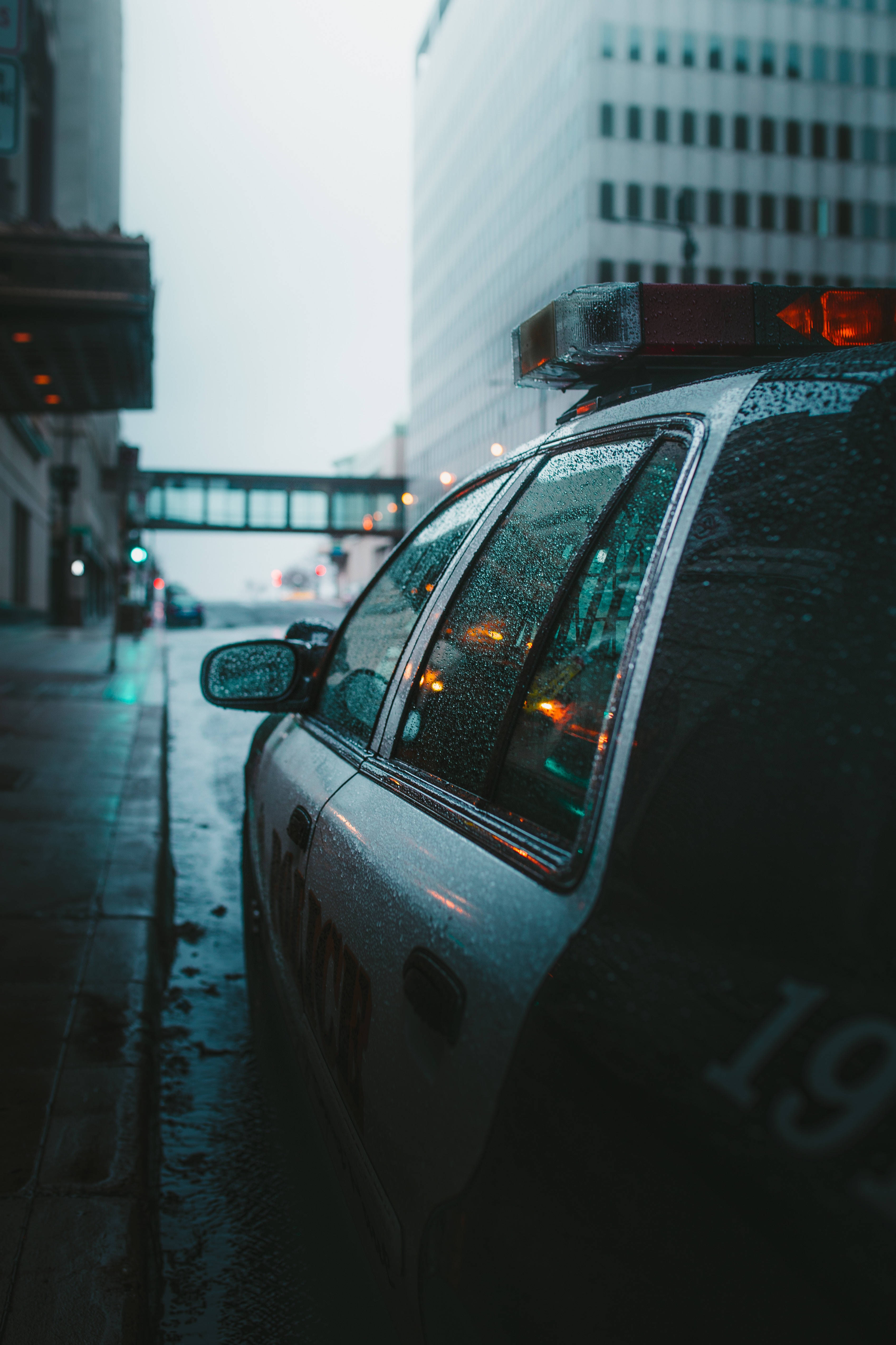 警察 桌布 壁紙 — 一輛被雨水淋濕的警車停泊在濕漉漉的城市街道上，背景是模糊的摩天大樓和發光的街燈。