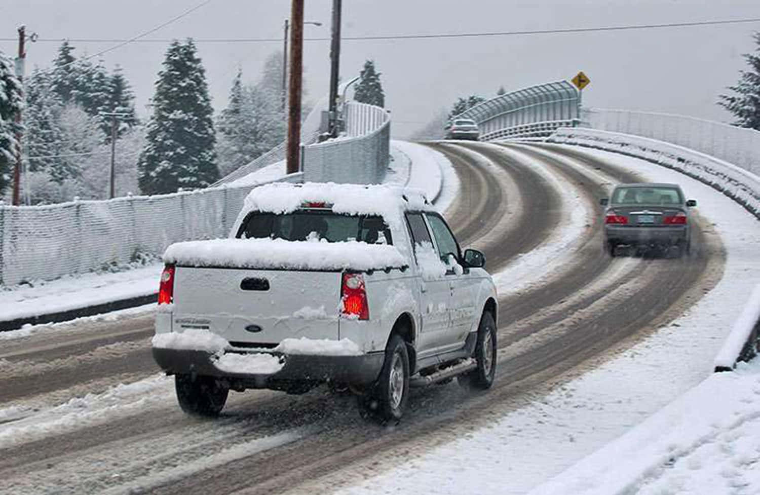 Winter Driving 桌布 壁紙 — 一輛被雪覆蓋的白色皮卡車在蜿蜒結冰的道路上行駛，道路兩旁是積雪的常青樹和護欄，天空多雲。