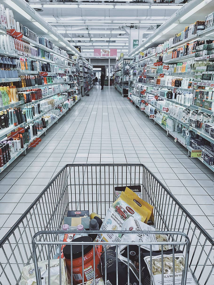 Shopping 桌布 壁紙 — 一輛裝滿雜貨的購物車放在光線充足的超市通道中央，通道兩旁擺滿個人護理產品的貨架。