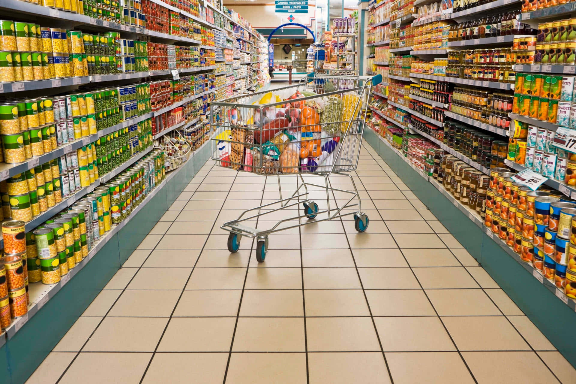 Grocery Store 桌布 壁紙 — 一輛裝滿雜貨的購物車放在光線明亮的超市走道中央，走道兩旁貨架陳列整齊。