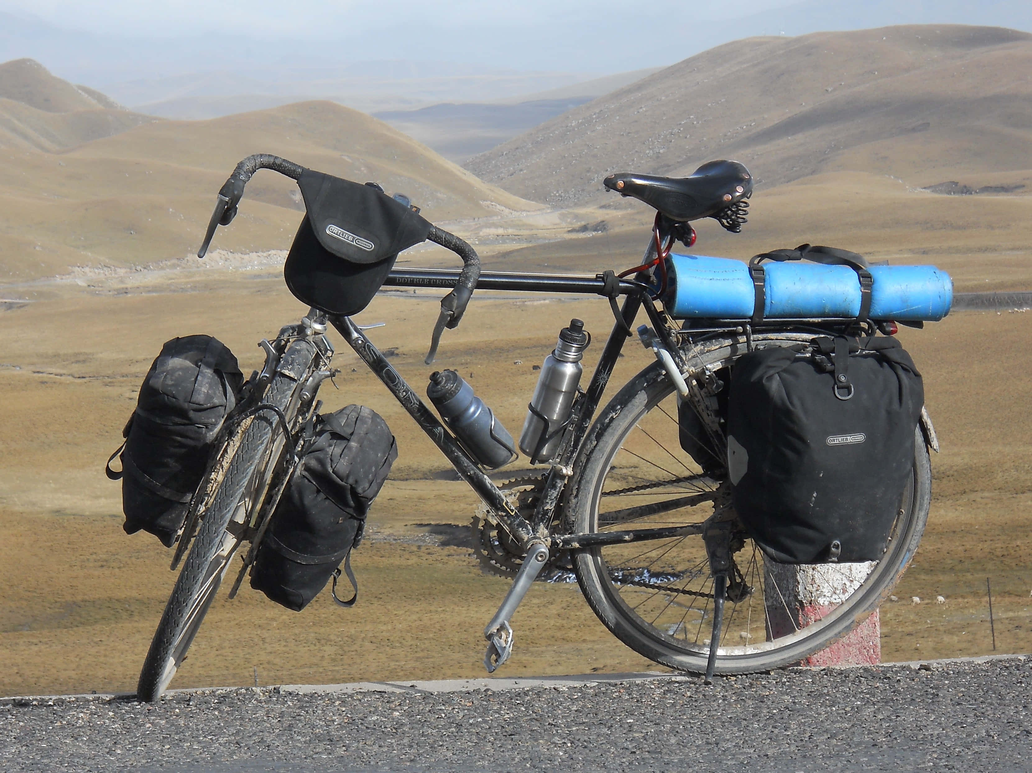 Bicycle 桌布 壁紙 — 一輛裝載著裝備的堅固旅行自行車在路邊休息，背景是朦朧天空下起伏的金色山丘。