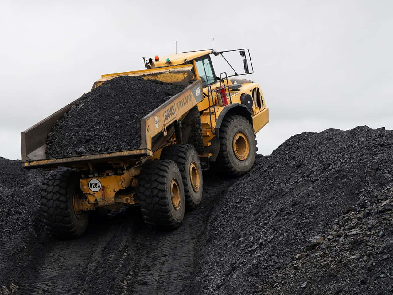 Coal Mining 桌布 壁紙 — 一輛裝載黑色煤炭的巨大黃色富豪自卸車在陰天爬升陡峭的煤堆。