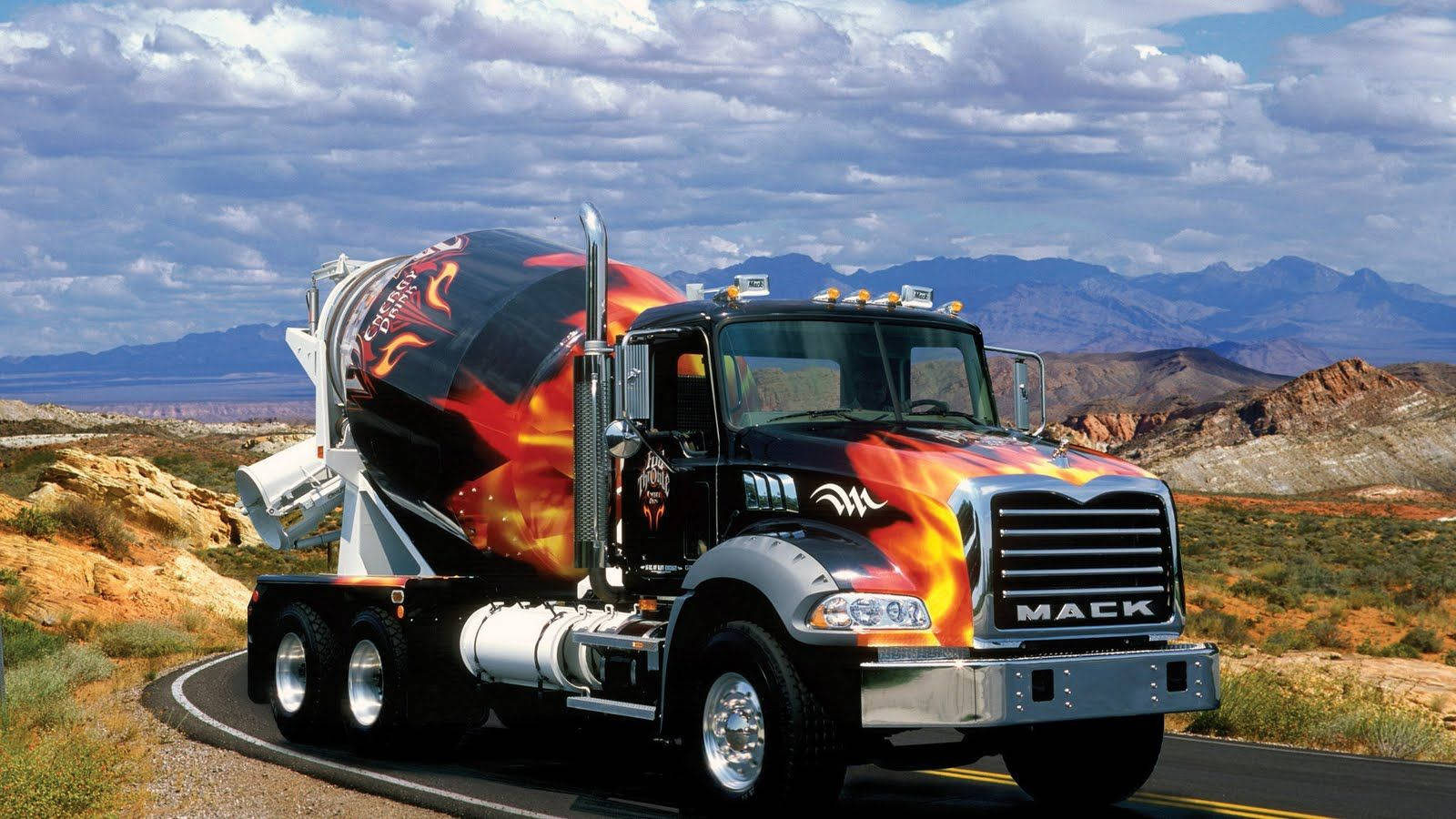 Trucks 桌布 壁紙 — 一輛裝飾著火焰圖案的Mack水泥車在雲層密布的天空下，行駛於有山脈的沙漠高速公路上。
