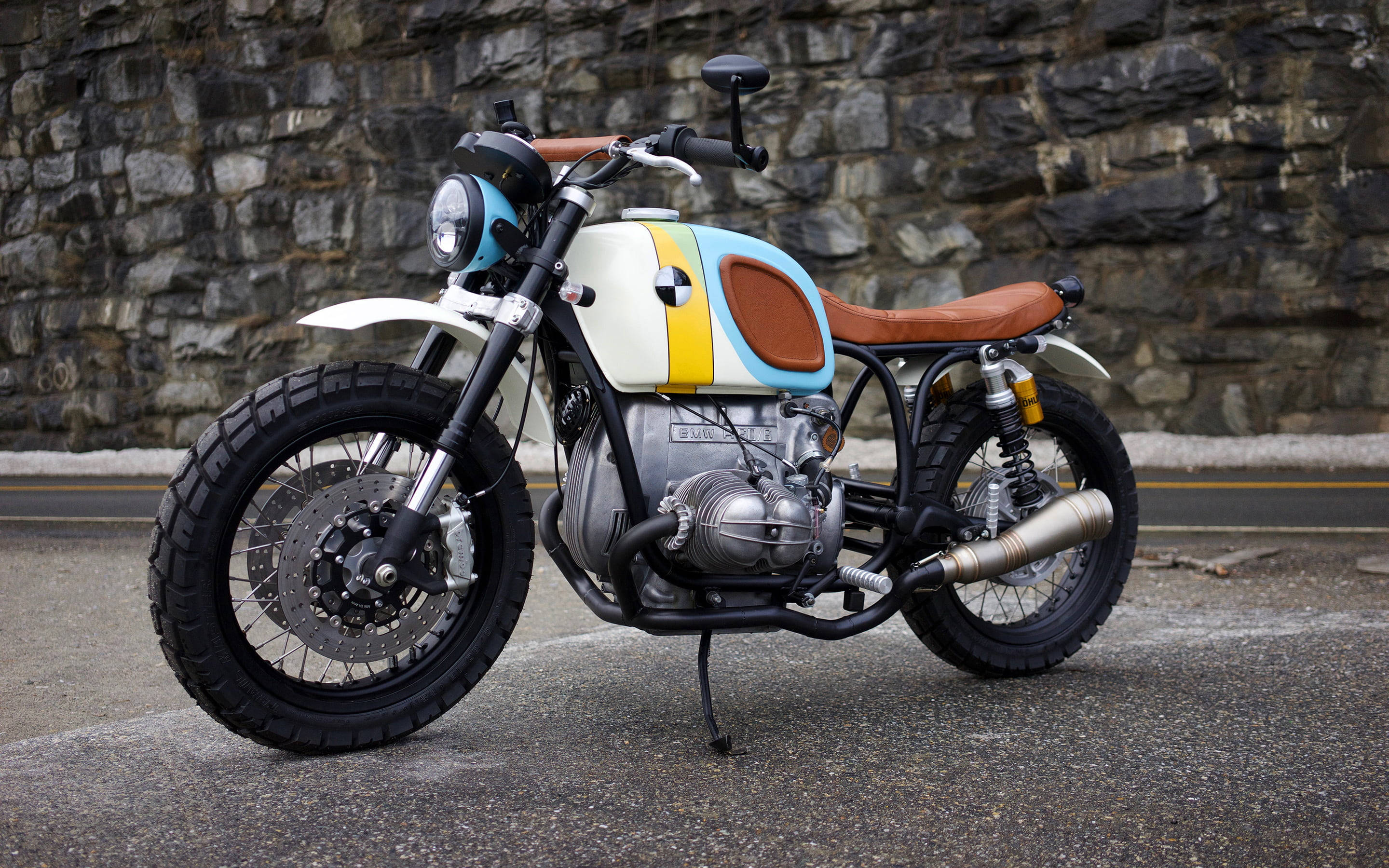 Bobber 桌布 壁紙 — 一輛訂製的咖啡賽車摩托車，帶有藍、黃、白色塗裝和棕色皮革座椅，停在柏油路面上，靠著石牆。