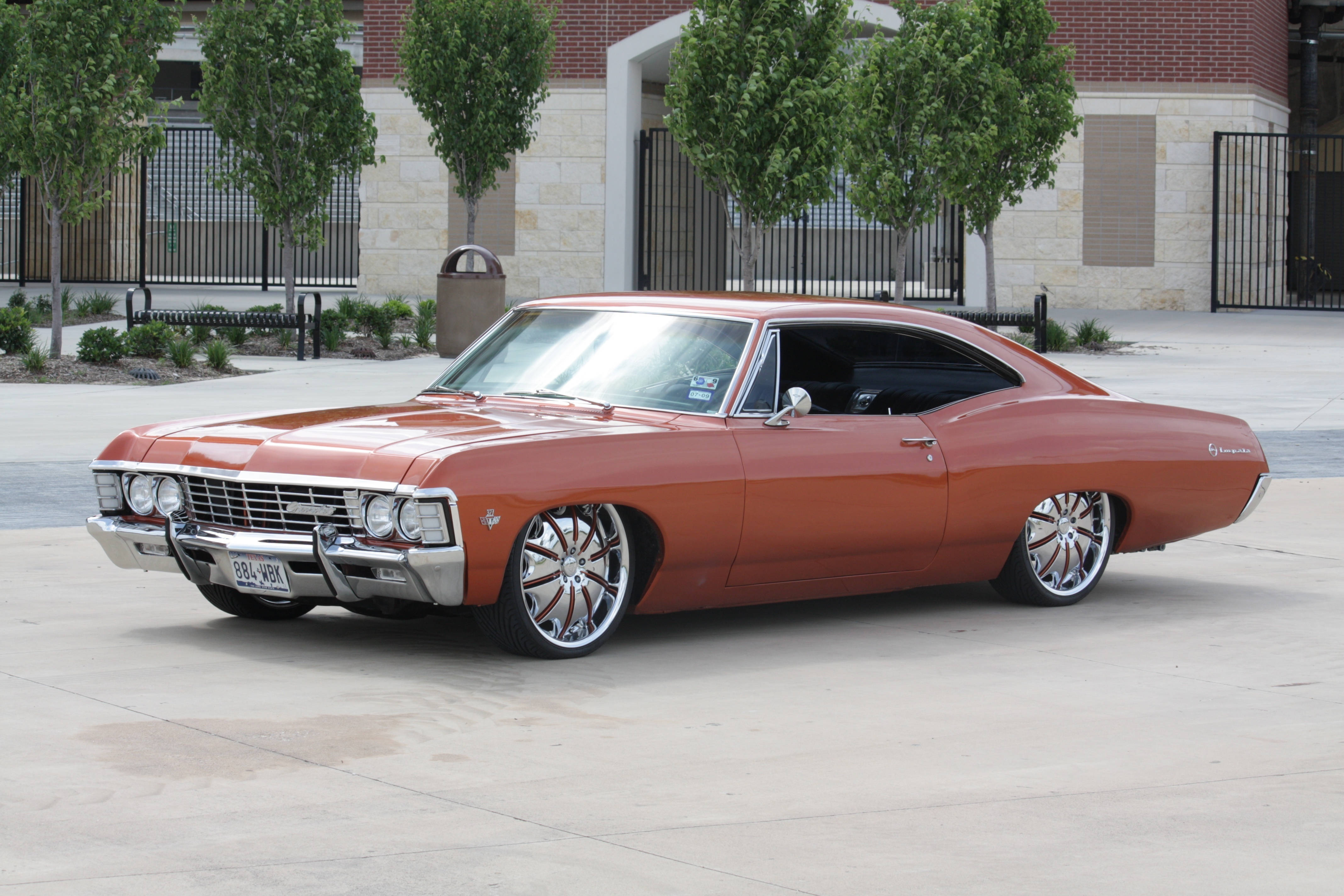 chevrolet impala 1967 桌布 壁紙 — 一輛訂製的橙色經典肌肉車，配備鍍鉻輪圈，停在混凝土停車場上，前方是磚砌建築和綠樹。